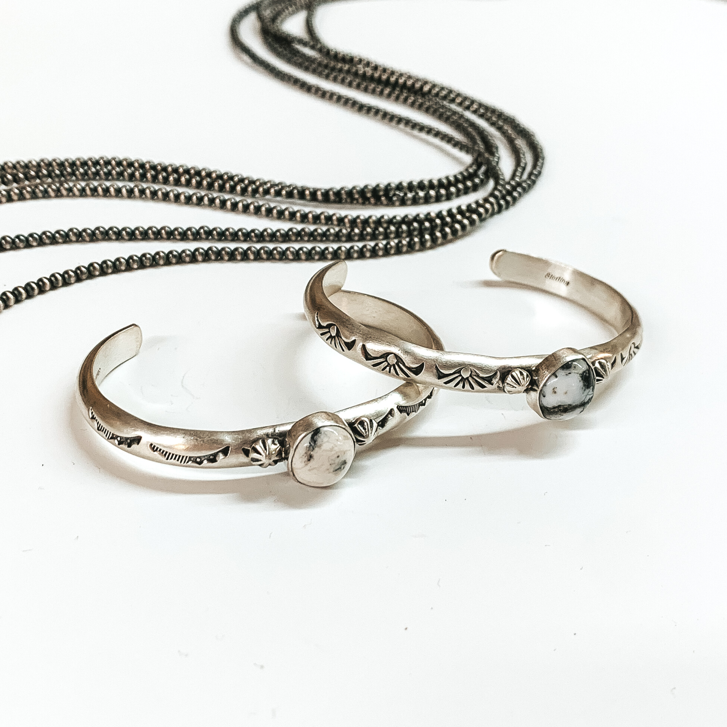 Two silver bangle bracelet with center white buffalo stones. These bangles also include engraved details. These bracelets are pictured on a white background with silver beads at the top of the picture. 
