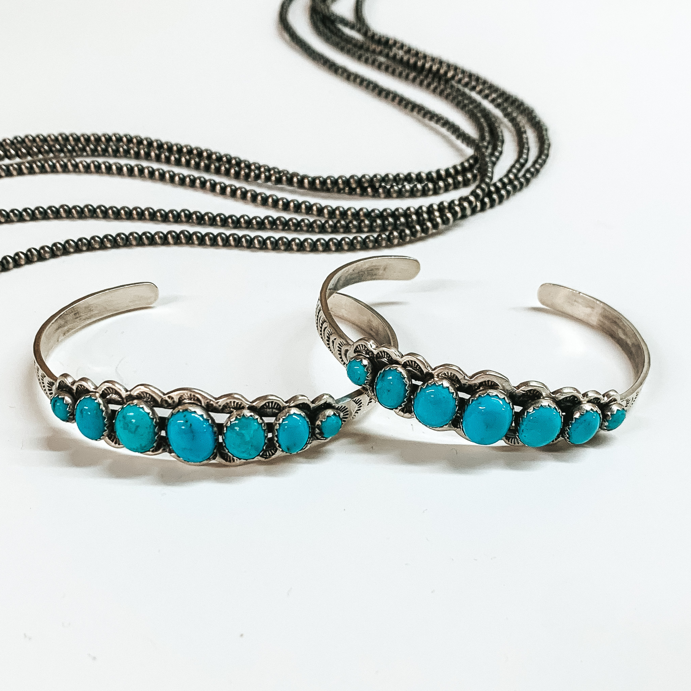 Two silver cuff bracelets with seven turquoise stones. These stones are arranged with the biggest in the middle and then they get gradually smalle on each side. These bracelets are pictured on a white background with silver beads at the top. 