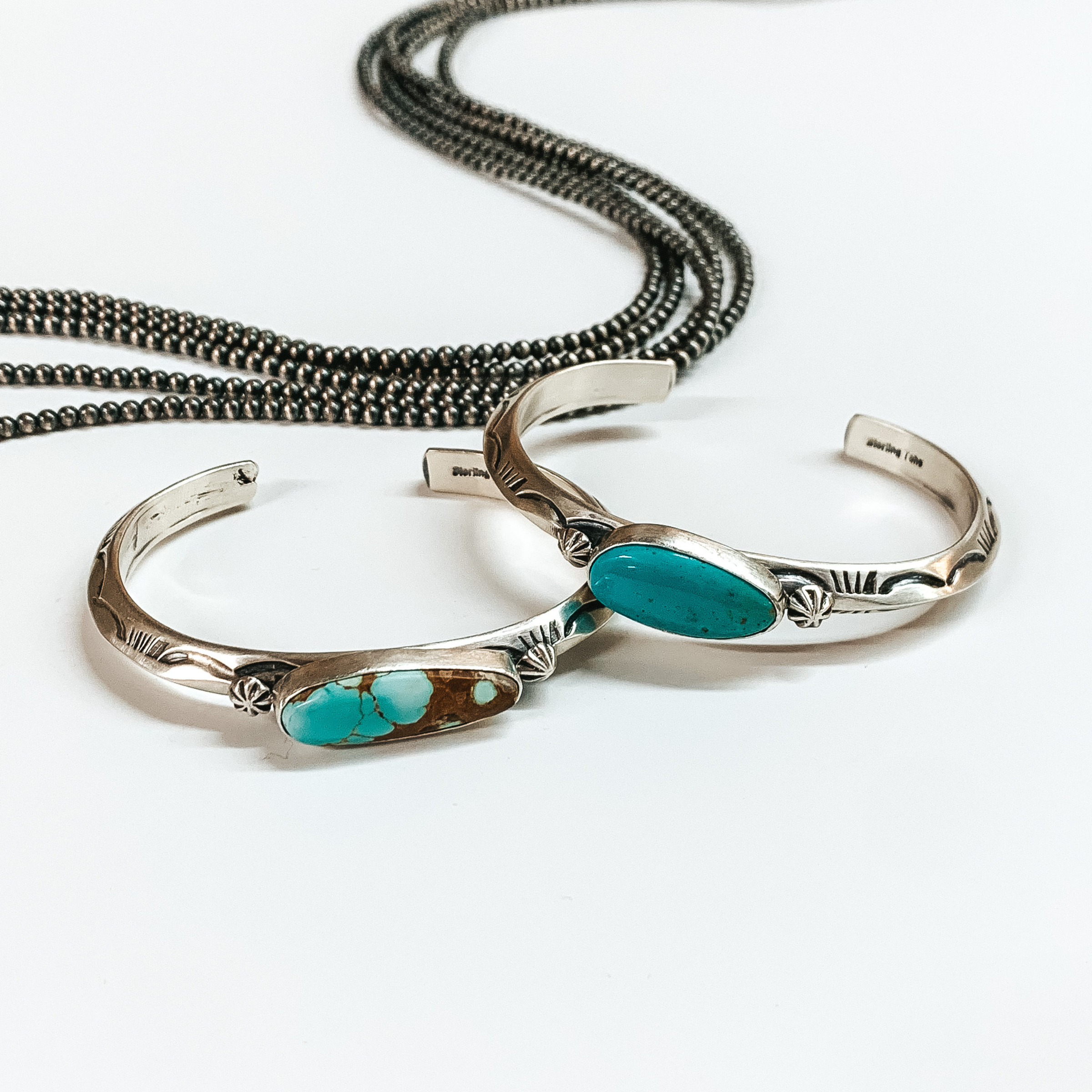 Two silver cuff bracelets with engraved details and a center turquoise stone. These bracelets are pictured on a white background with silver beads at the top. 