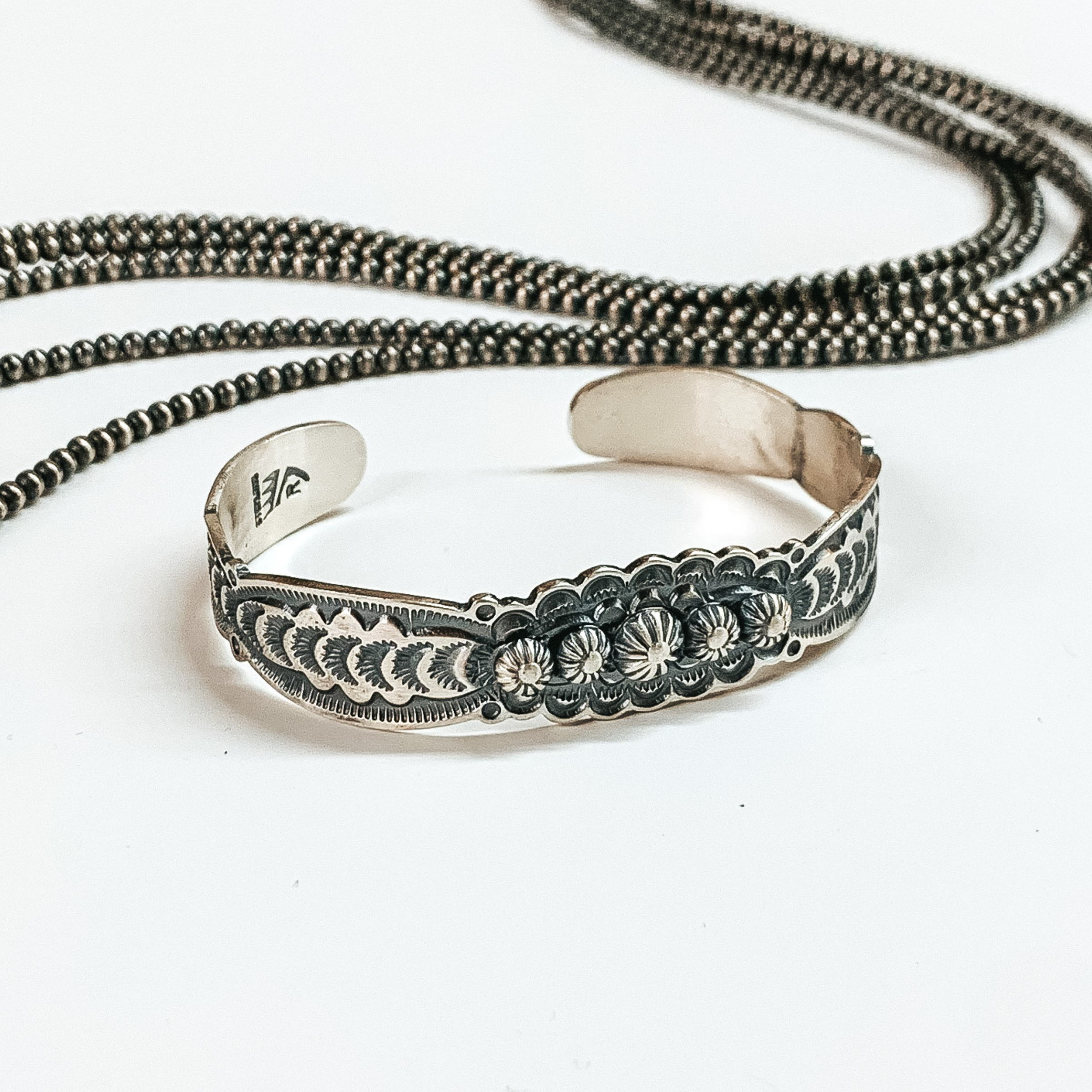 Silver cuff bracelet with a tooled design. These bracelets are pictured on a white background with silver beads at the top. 