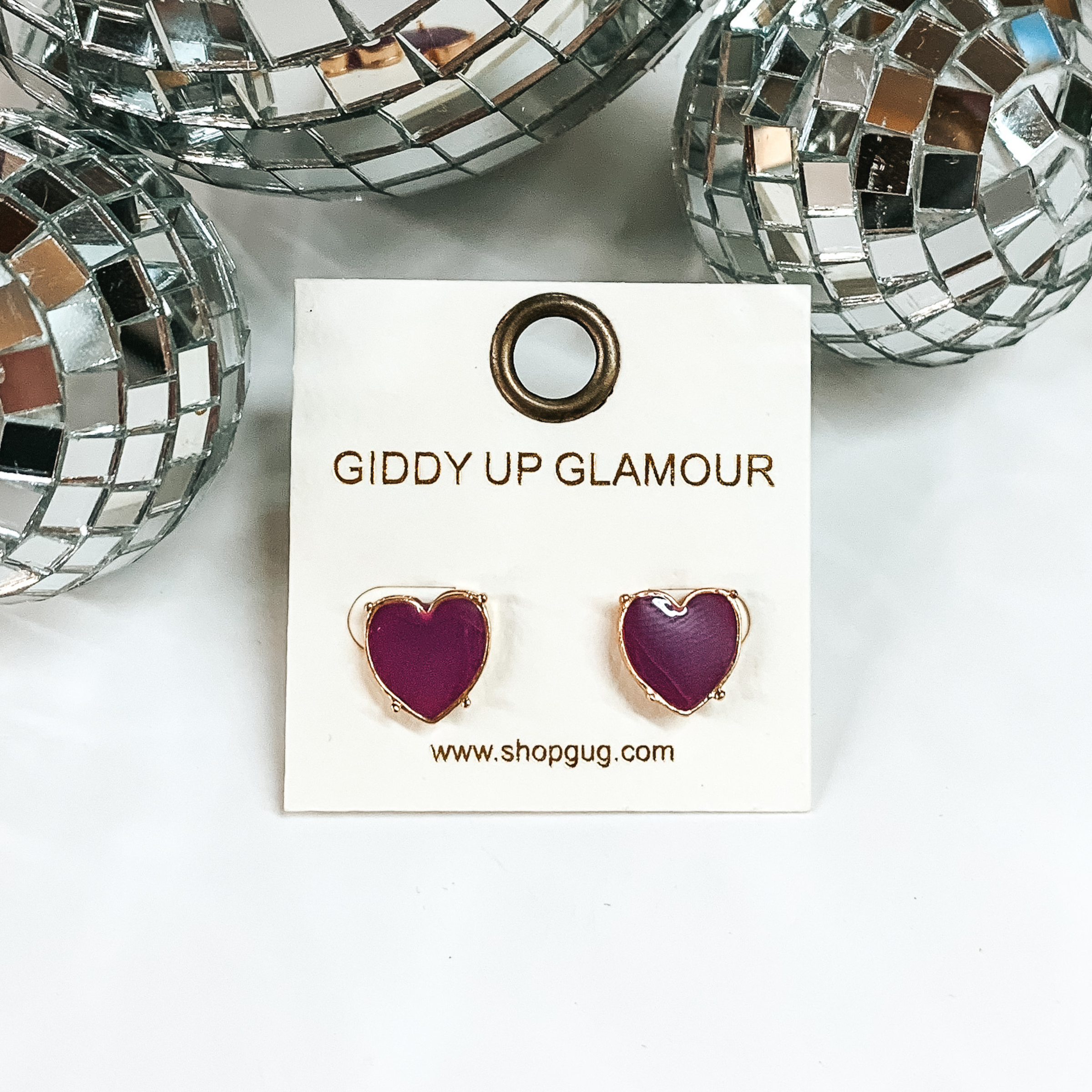 Dark purple heart stud earrings with a gold outline. These earrings are pictured on a white background with disco balls at the top of the picture. 
