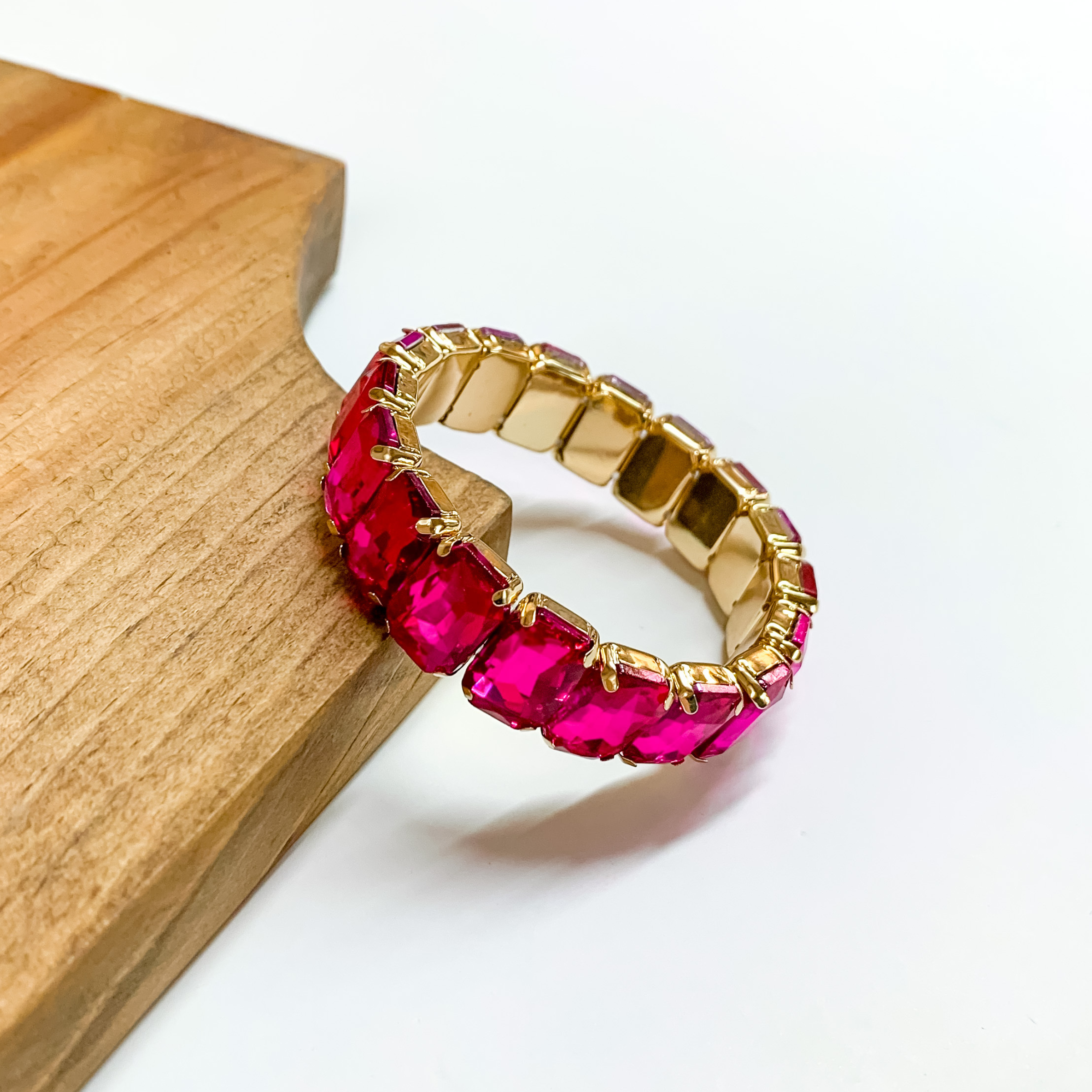 Hot pink rectangle crystal bracelet with a gold setting. This bracelet is pictured partially laying on a wood block on a white background. 