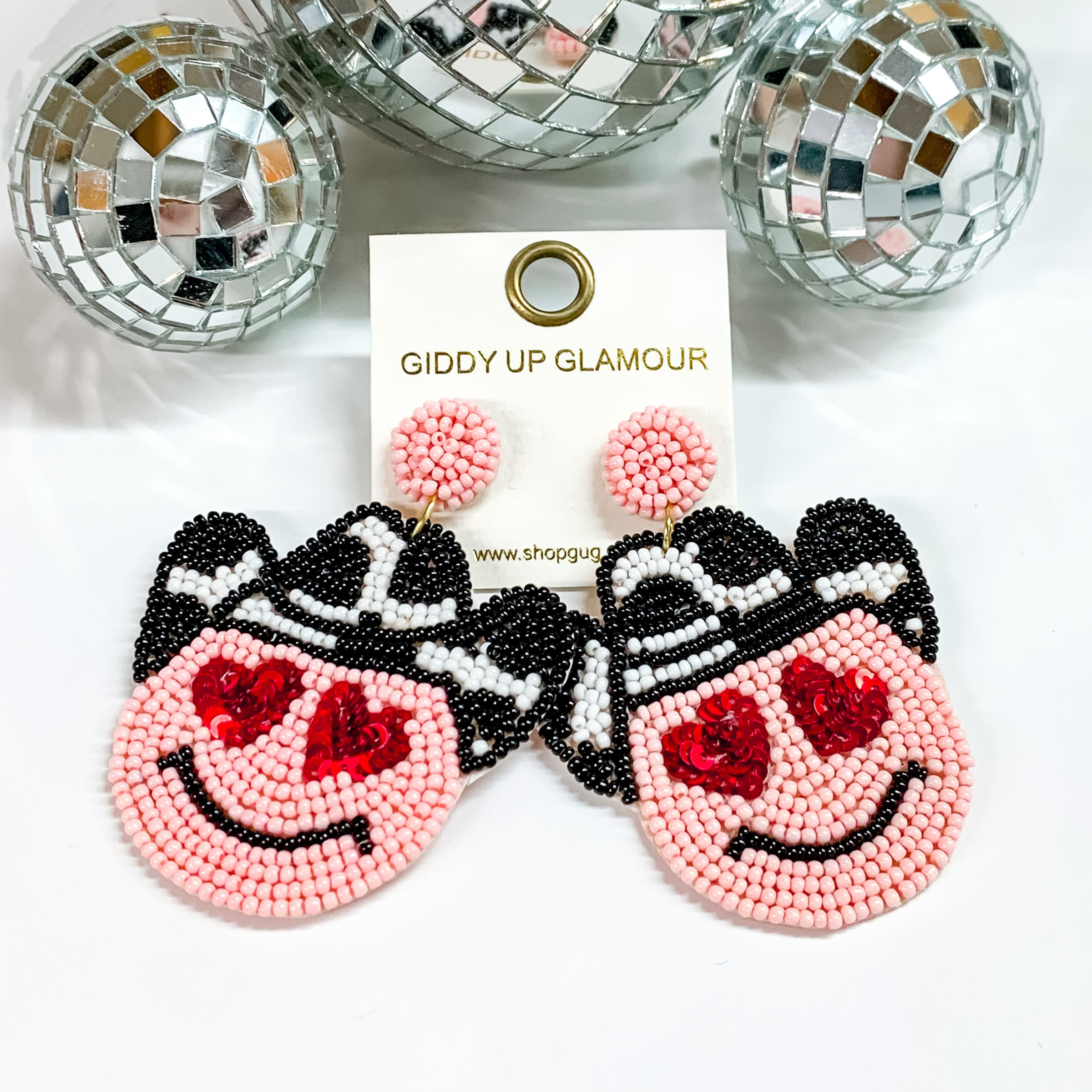 Pink smiley face drop earrings that include and black and white cow print cowboy hat and red heart eyes. These earrings are pictured on a white background with disco balls at the top of the picture. 