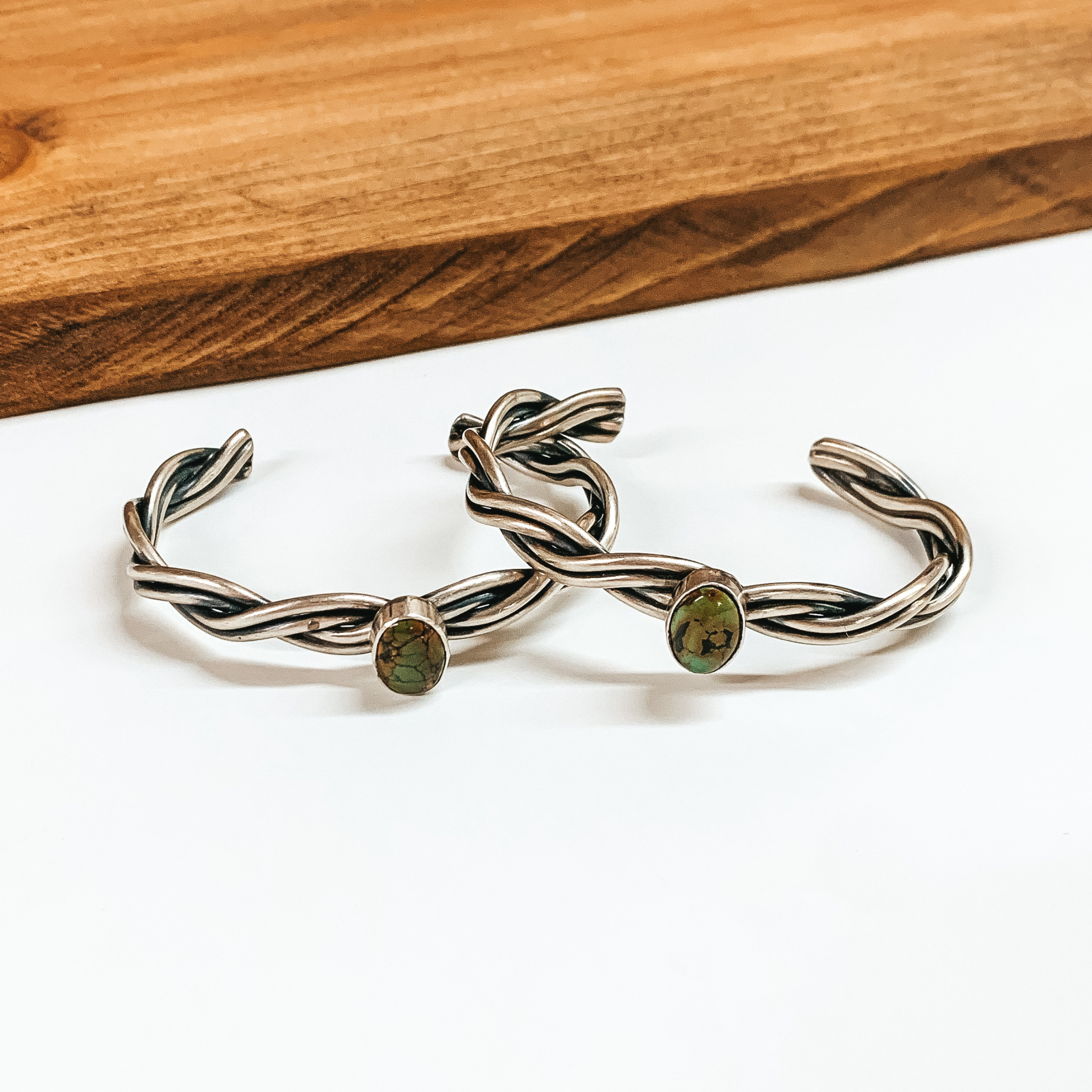 Two silver, rope cuff bracelets with oval center turquoise stones. These bracelets are pictured in front of a wood block on a white background. 