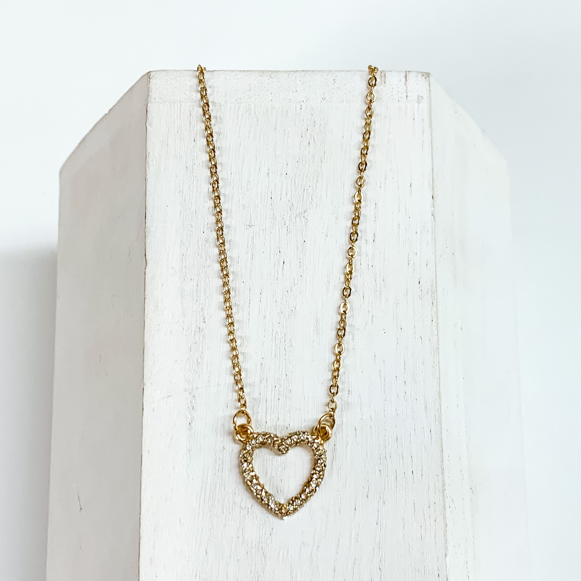 Gold chain necklace with a gold heart pendant that is encrusted with clear crystals. This necklace is laying on a white block on a white background. 