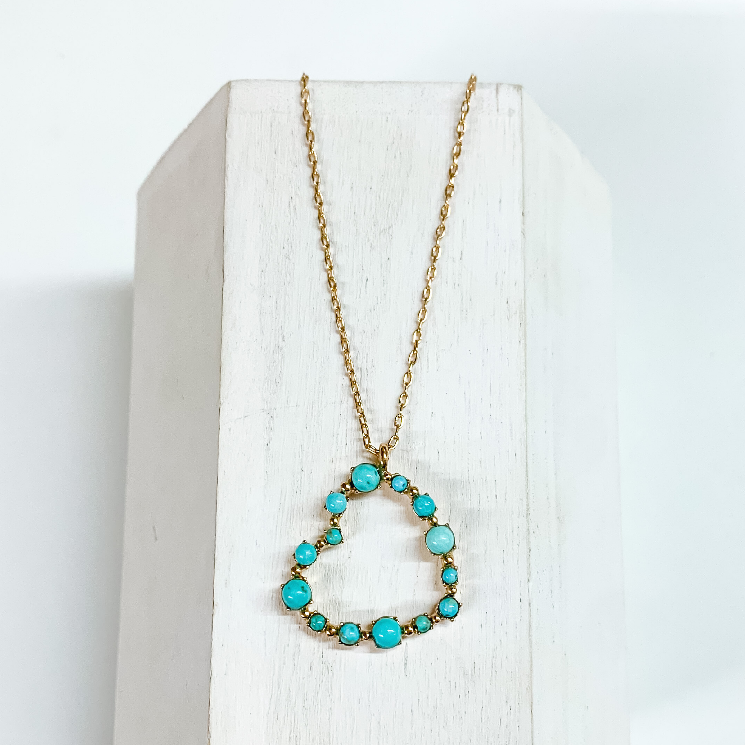 Gold chain necklace with a gold heart pendant that has turquoise stones outlining it. This necklace is pictured on a white block on a white background. 