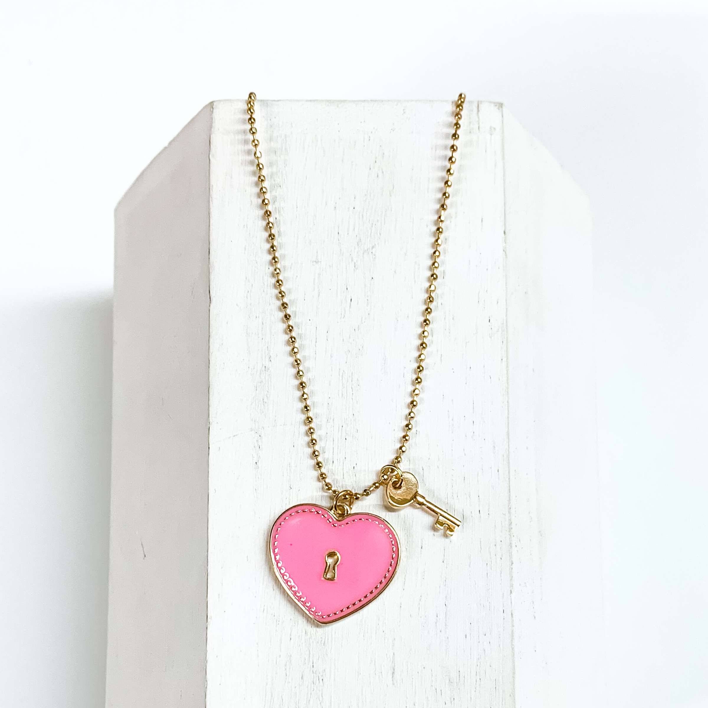 Gold, ball chain necklace with a light pink heart pendant and gold key charm. This necklace is pictured on a white block on a white background. 
