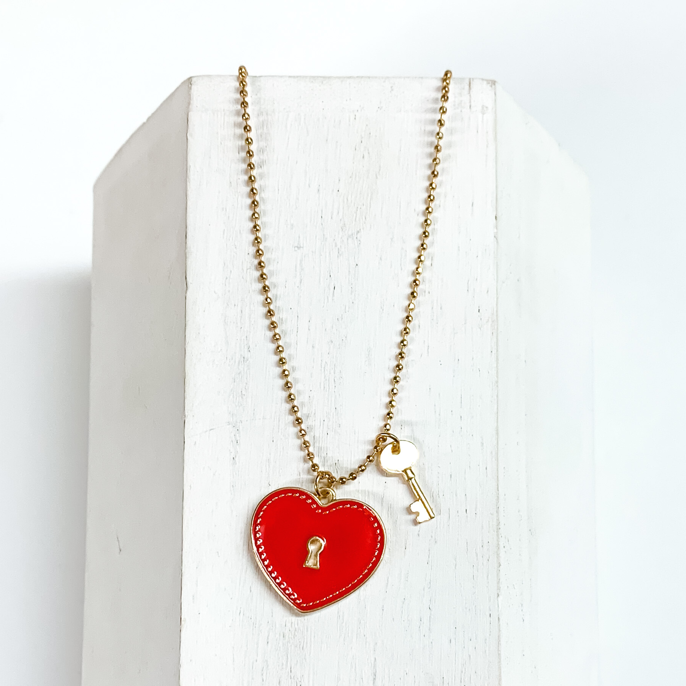 Gold, ball chain necklace with a red heart pendant and gold key charm. This necklace is pictured on a white block on a white background.