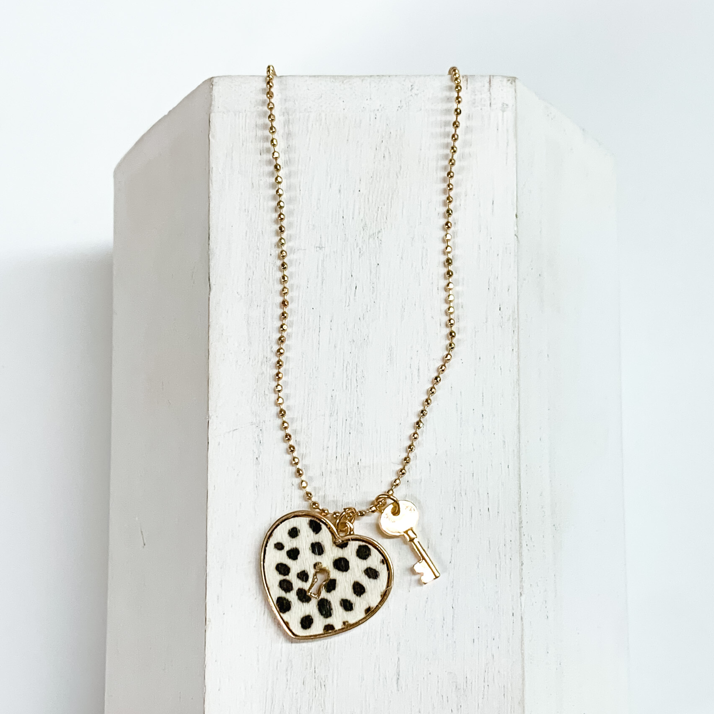 Gold, ball chain necklace with a white dotted heart pendant and gold key charm. This necklace is pictured on a white block on a white background.