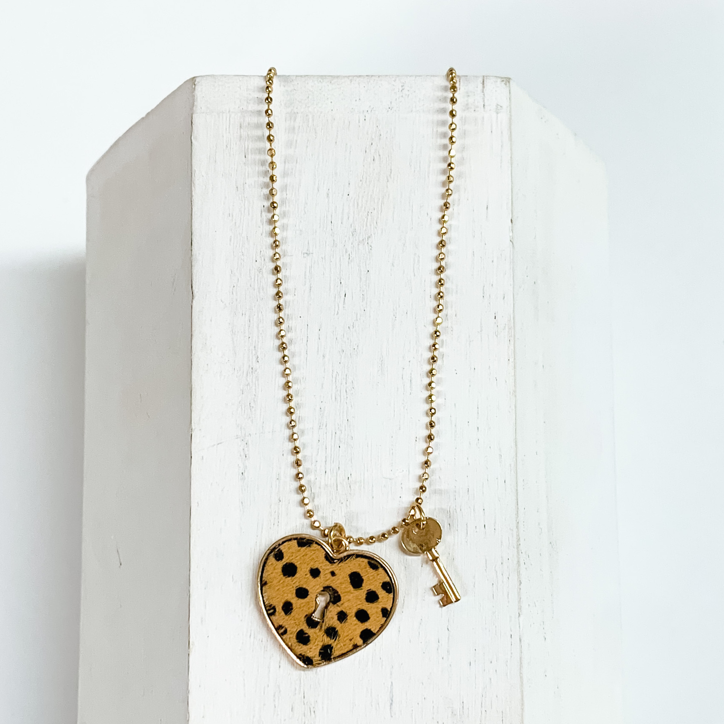 Gold, ball chain necklace with a brown dotted heart pendant and gold key charm. This necklace is pictured on a white block on a white background.
