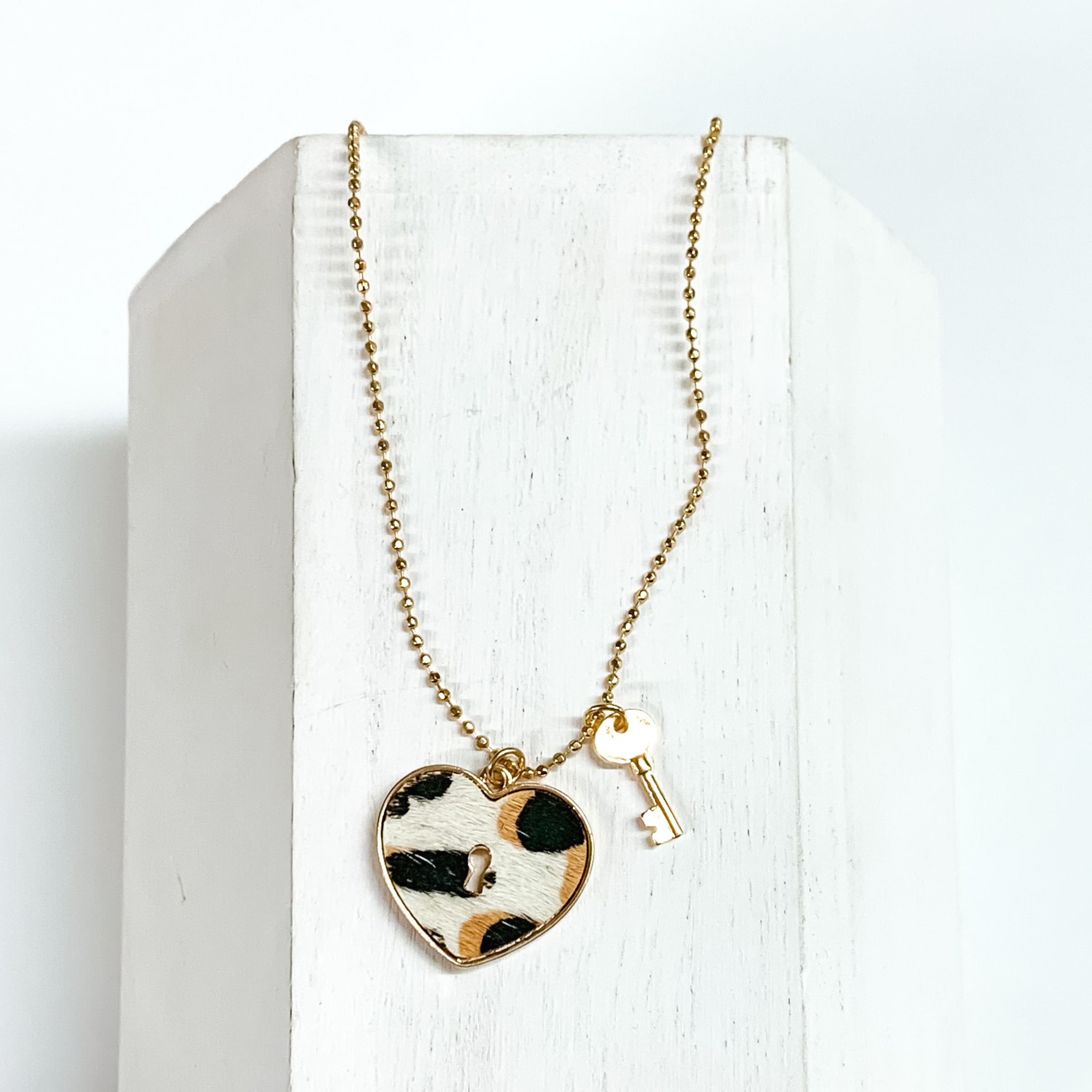 Gold, ball chain necklace with a white leopard print heart pendant and gold key charm. This necklace is pictured on a white block on a white background.