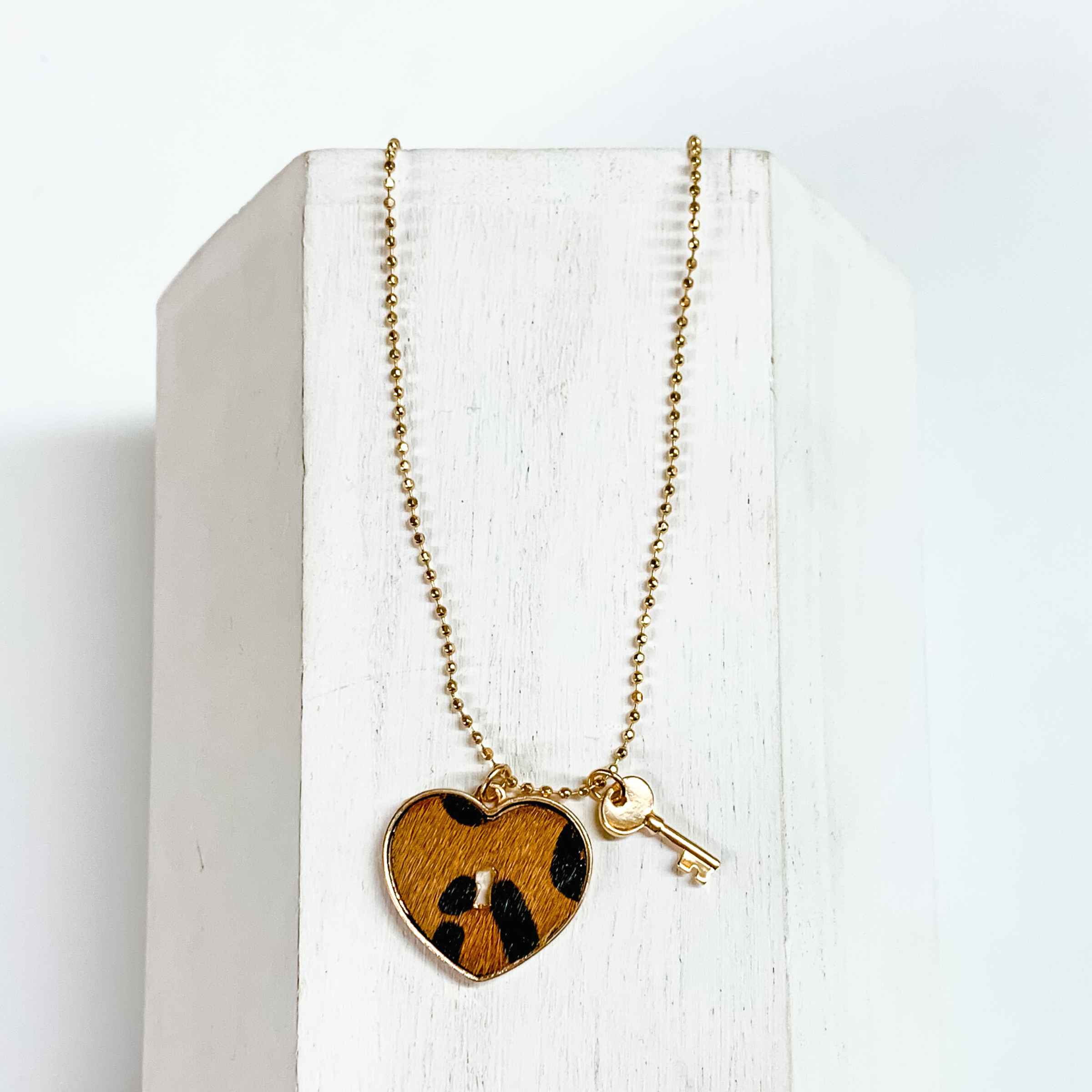 Gold, ball chain necklace with a brown leopard print heart pendant and gold key charm. This necklace is pictured on a white block on a white background.
