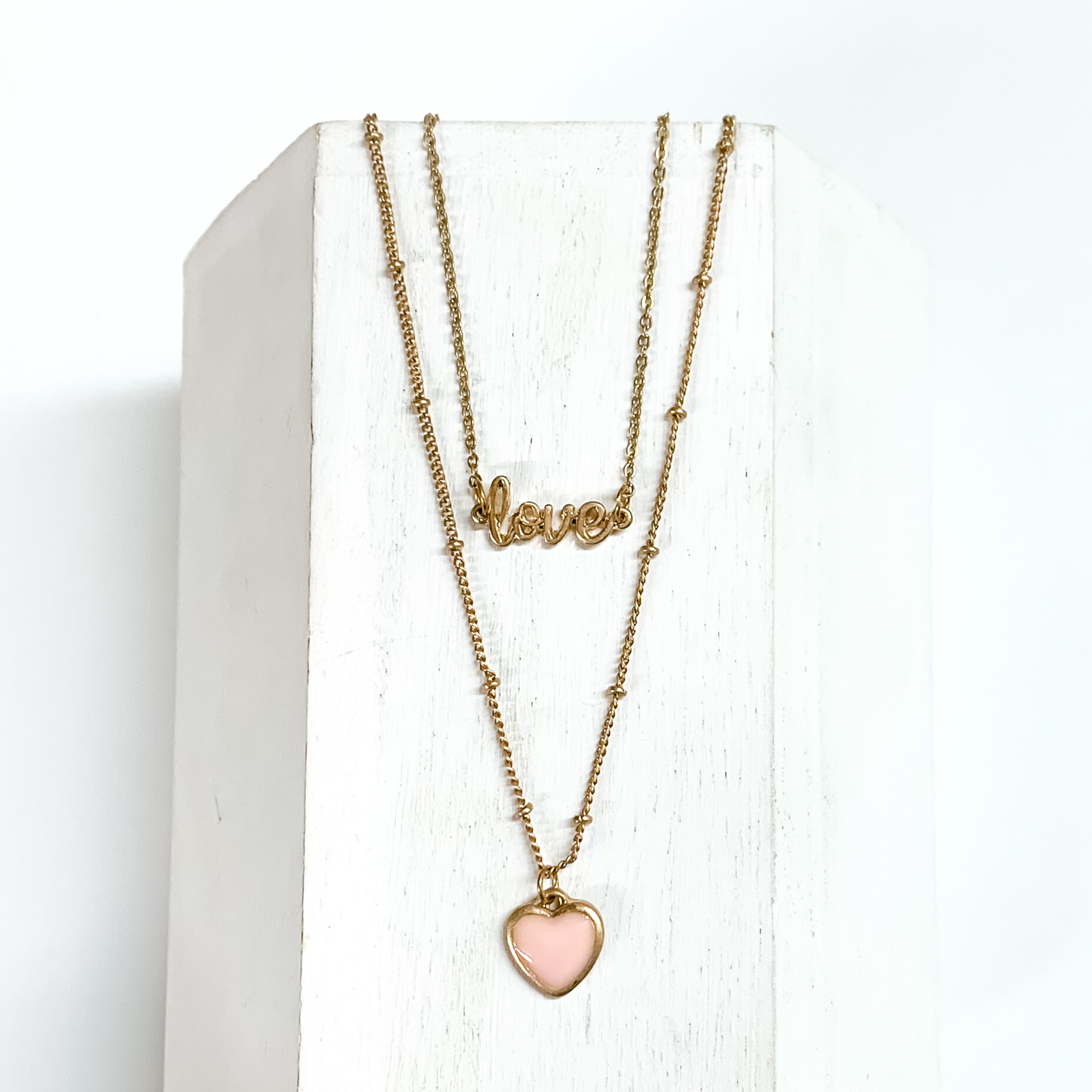 Two stranded gold chained necklace. The shorter strand has a gold pendant that spells out "LOVE" and the longer strand has a small pink heart charm. This necklace is pictured laying on a white block on a white background.