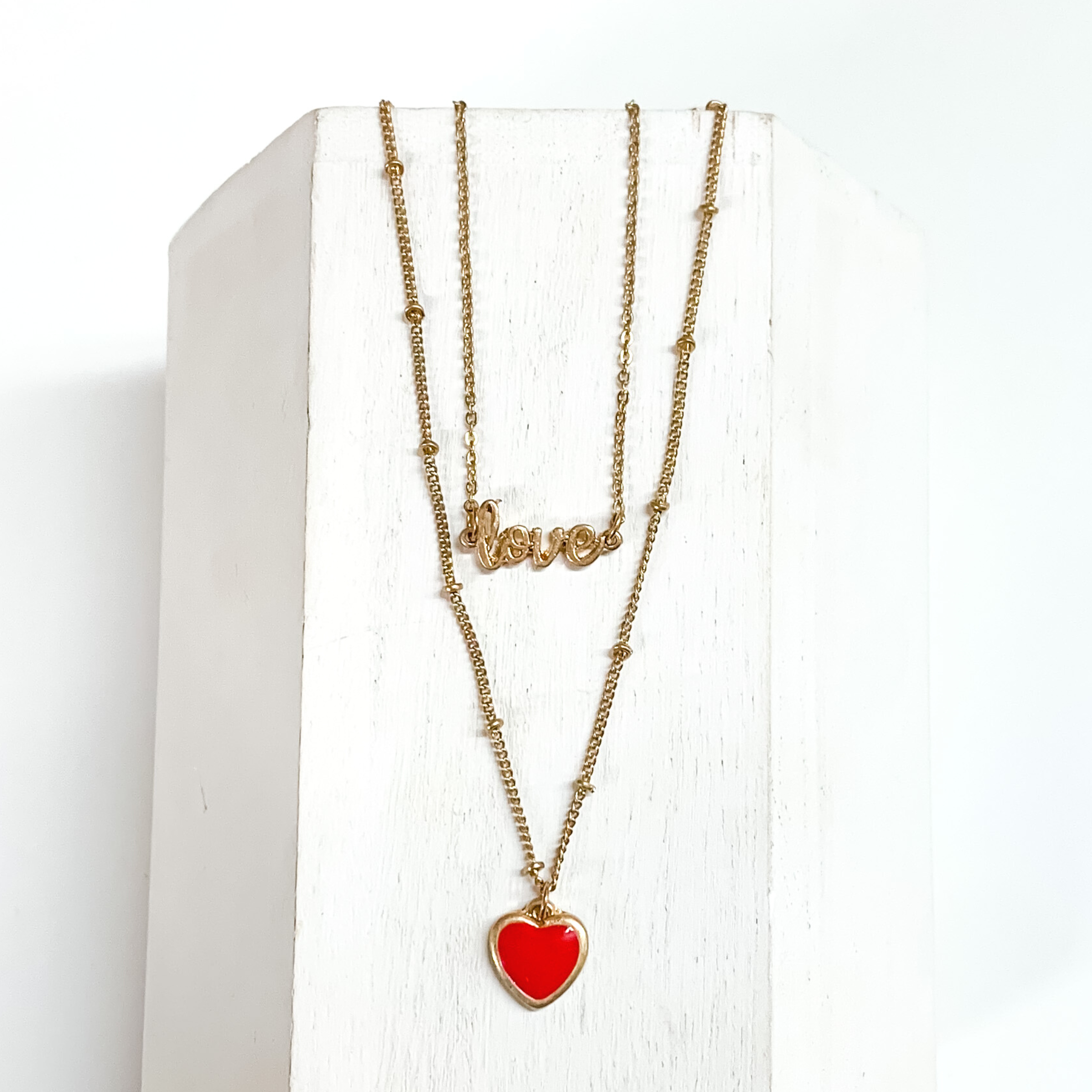 Two stranded gold chained necklace. The shorter strand has a gold pendant that spells out "LOVE" and the longer strand has a small red heart charm. This necklace is pictured laying on a white block on a white background.