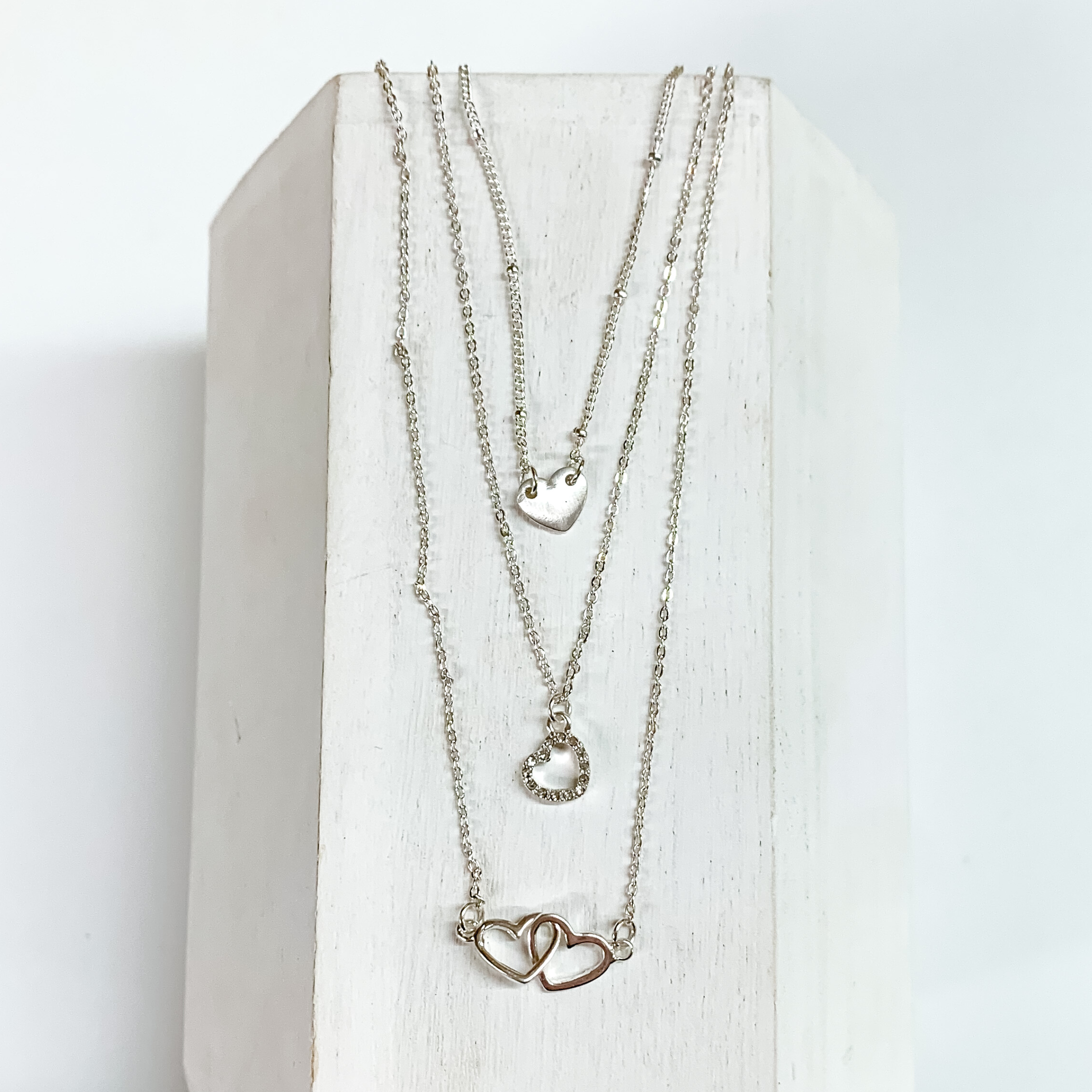 Three silver chain strands each with a heart pendant. The shortest strand has a plain silver heart, medium chain has a heart outline pendant, and the longest strand has two interlocking hearts pendant. This necklace is pictured on a white block on a white background. 