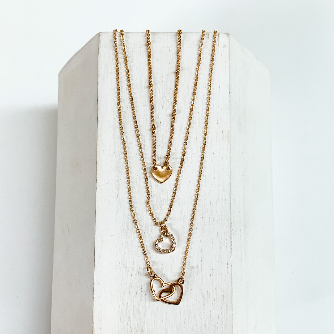Three gold chain strands each with a heart pendant. The shortest strand has a plain gold heart, medium chain has a heart outline pendant, and the longest strand has two interlocking hearts pendant. This necklace is pictured on a white block on a white background. 