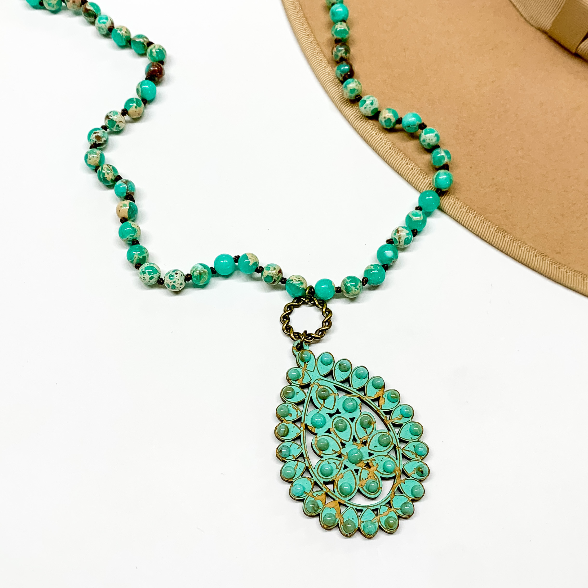 Turquoise beaded necklace with a turquoise teardrop pendant connected to th necklace by a bronze circle. This necklace is pictured on a white background with a tan hat brim in the top right corner. 