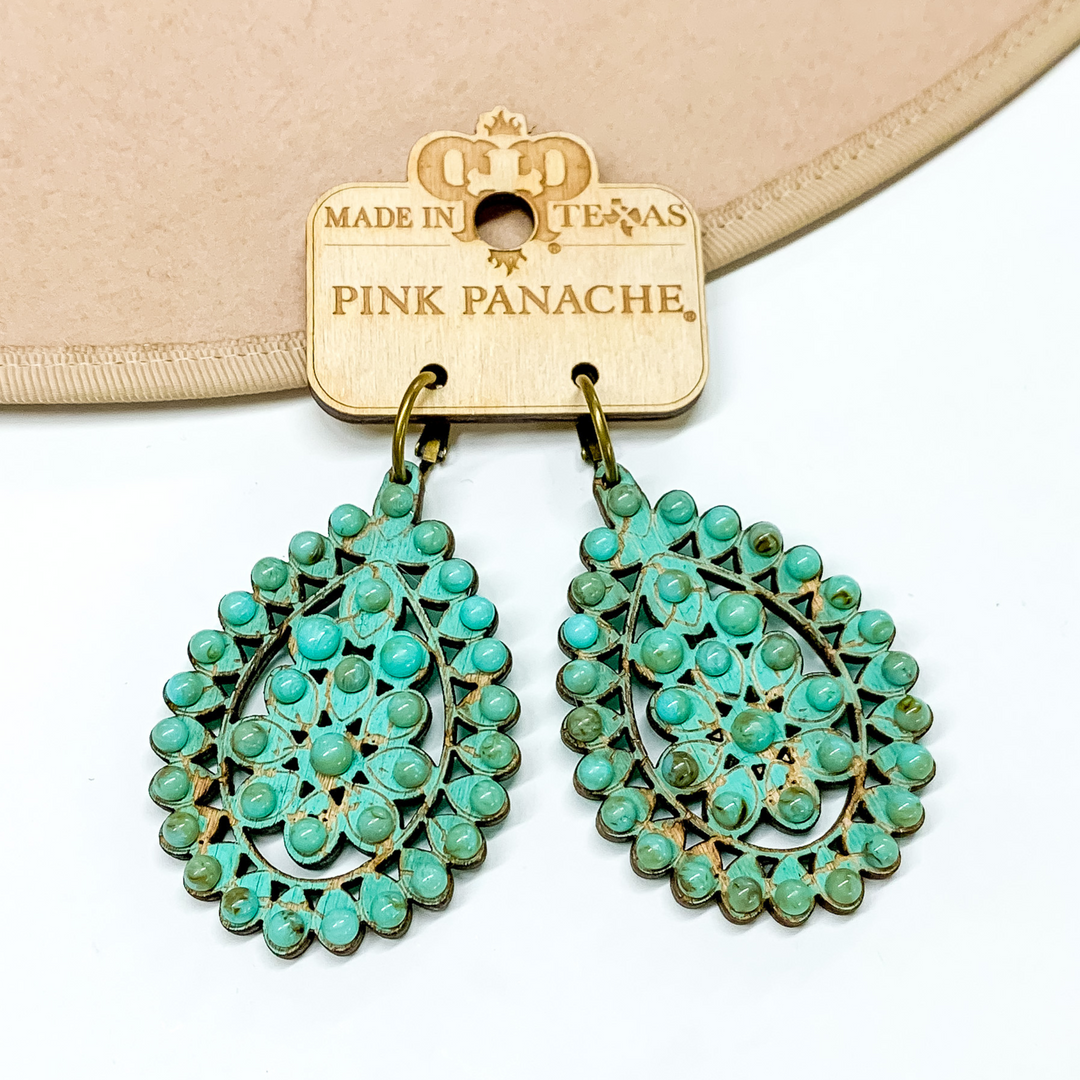 Bronze hoop earrings with turquoise, teardrop pendant with turquoise bead outline. These earrings are pictured partially laying on a tan hat brim on a white background. 