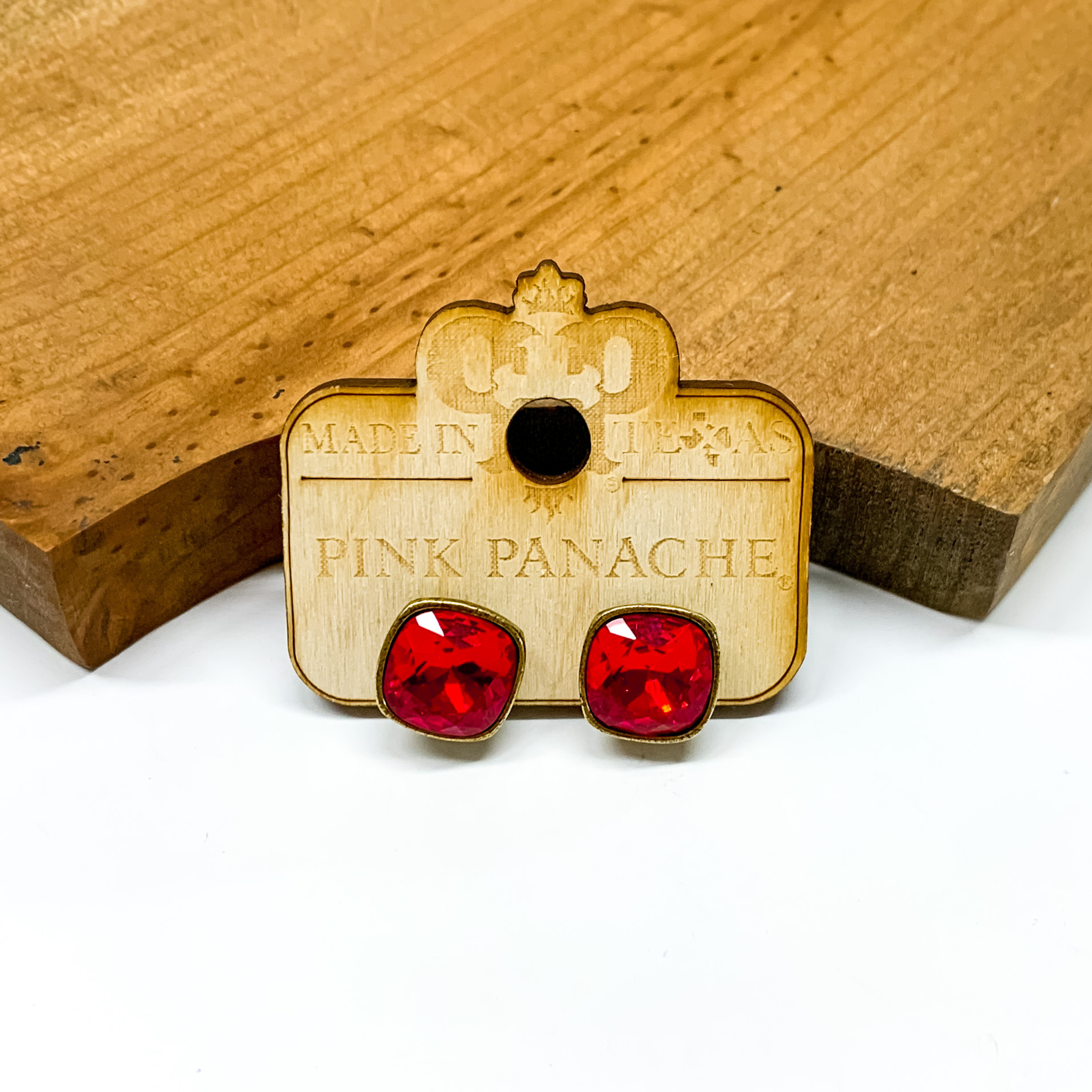 Square red crystal stud earrings pictured on a wood earrings holder. These earrings are pictured in front of a wood block on a white background. 