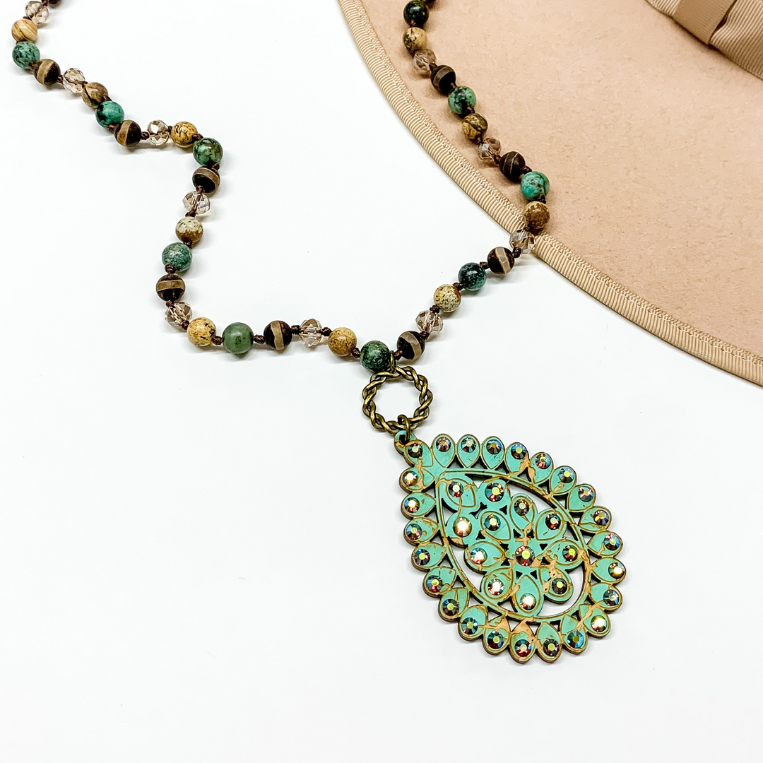 Brown mix beaded necklace with a turquoise teardrop pendant connected to the necklace by a bronze circle. The pendant has ab crystal outline. This necklace is pictured on a white background with a tan hat brim in the top right corner. 