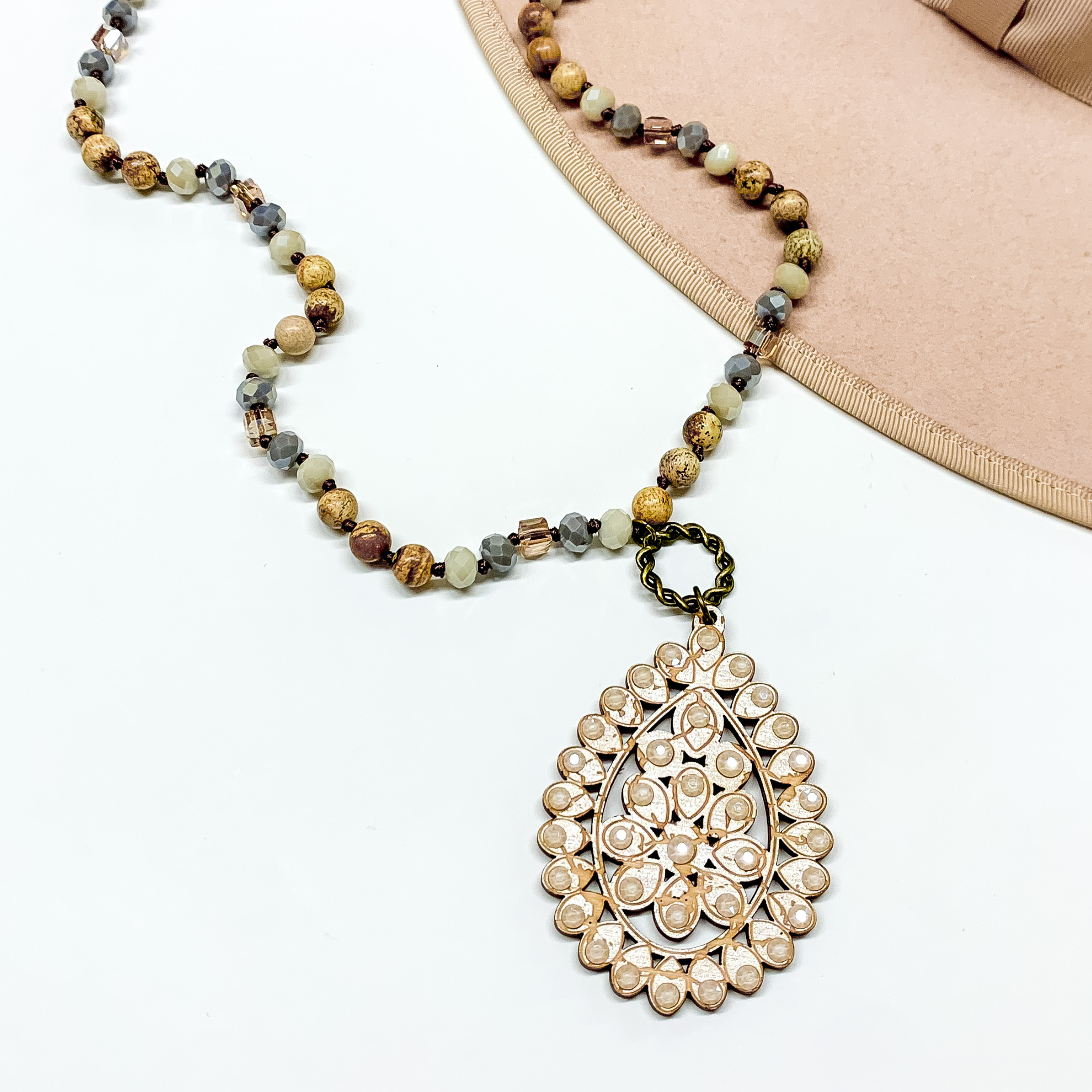 Brown mix beaded necklace with a tan teardrop pendant connected to the necklace by a bronze circle. The pendant has a candy ivory crystal outline. This necklace is pictured on a white background with a tan hat brim in the top right corner. 