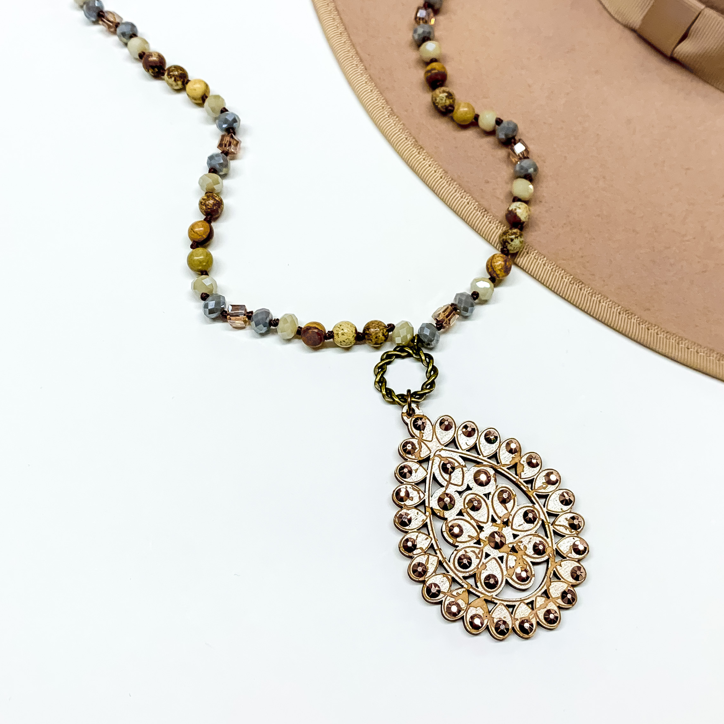 Brown mix beaded necklace with a tan teardrop pendant connected to the necklace by a bronze circle. The pendant has a rose gold crystal outline. This necklace is pictured on a white background with a tan hat brim in the top right corner. 
