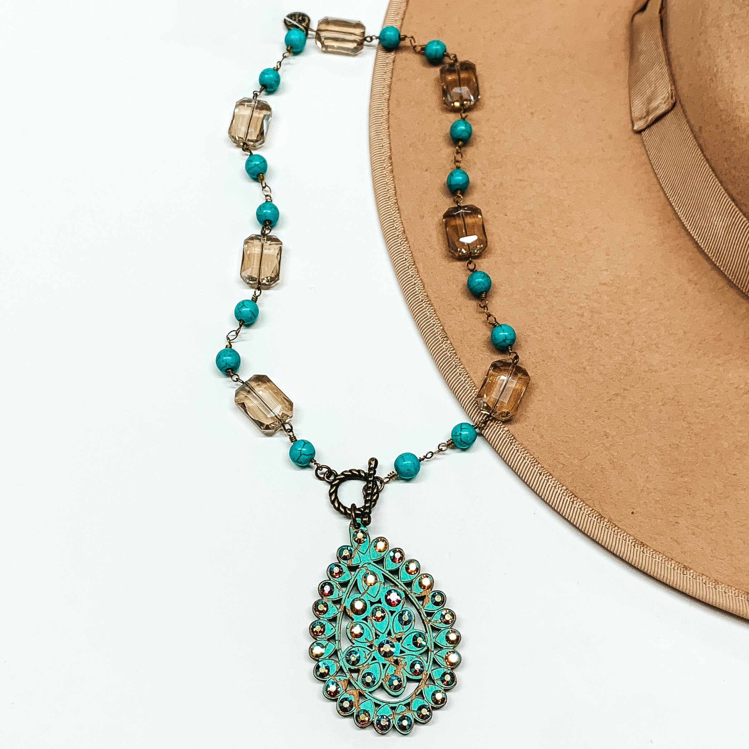 This necklace has round turquoise beads and rectangle light tan beads connected by bronze chains. This necklace also includes a toggle clasp that has has a turquoise teardrop pendant hat includes ab crystal outlines. 