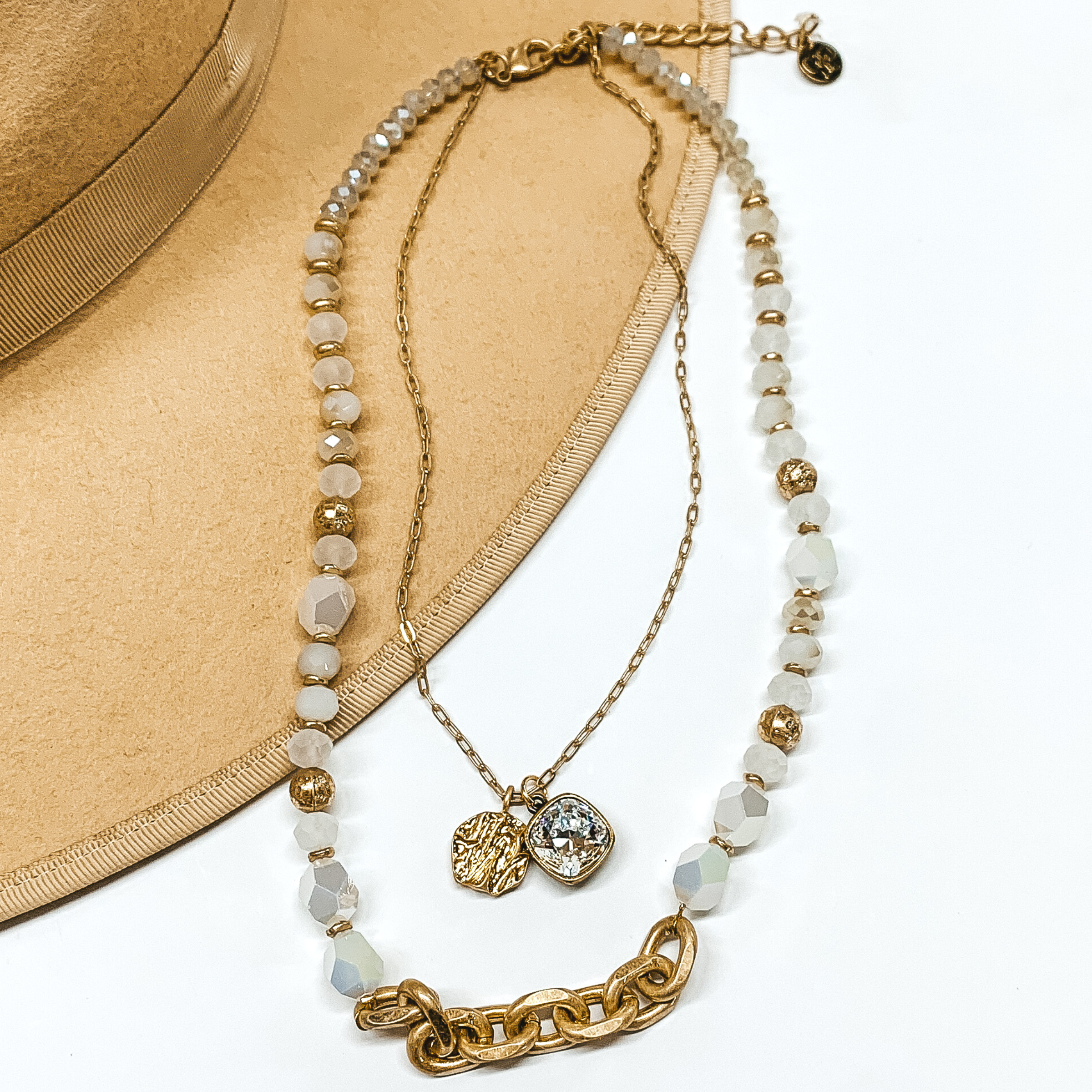 This is a two strand necklace that has one short dainty chain and a longer white crystal beaded strand with a gold chain segment. The shorter chain includes a gold coin charm and clear cushion cut crytal charm. This necklace is pictured partially laying on a tan brim on a white background. 
