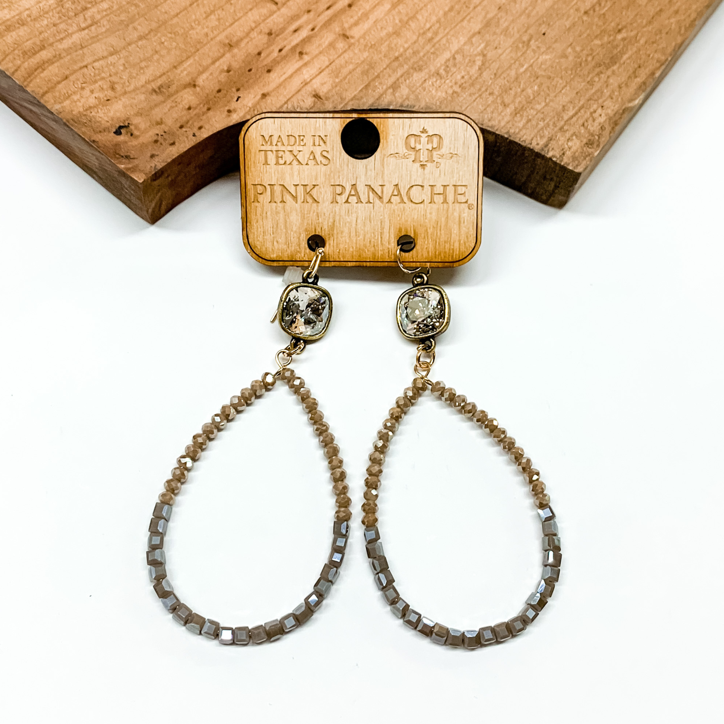 Brown and grey beaded teardrop earrings with a clear cushion cut connector. These earrings are pictured in front of a brown block on a white background. 