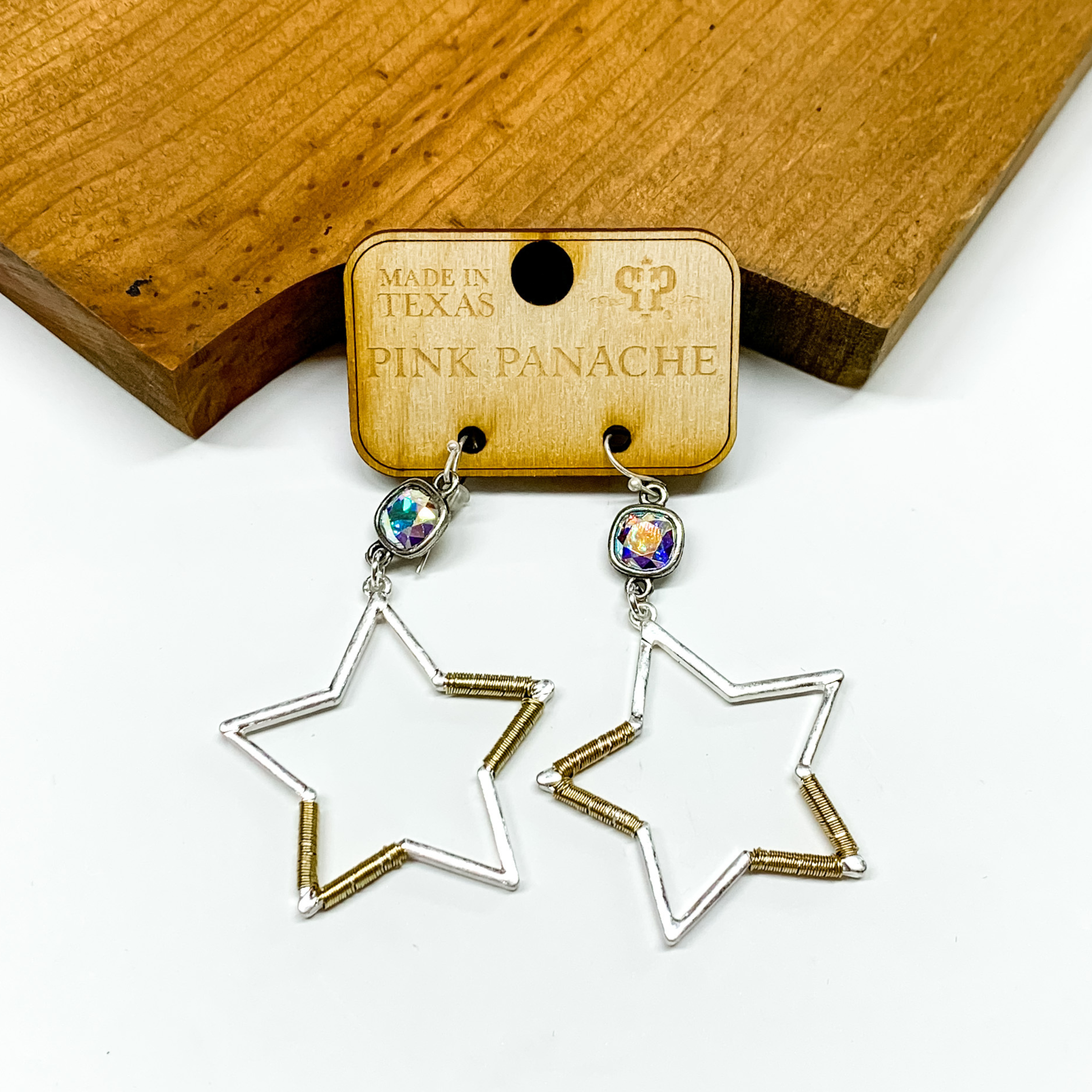 Silver and gold star earrings with an ab cushion cut crystal connectors. These earrings are pictured in front of a wood block on a white background. 