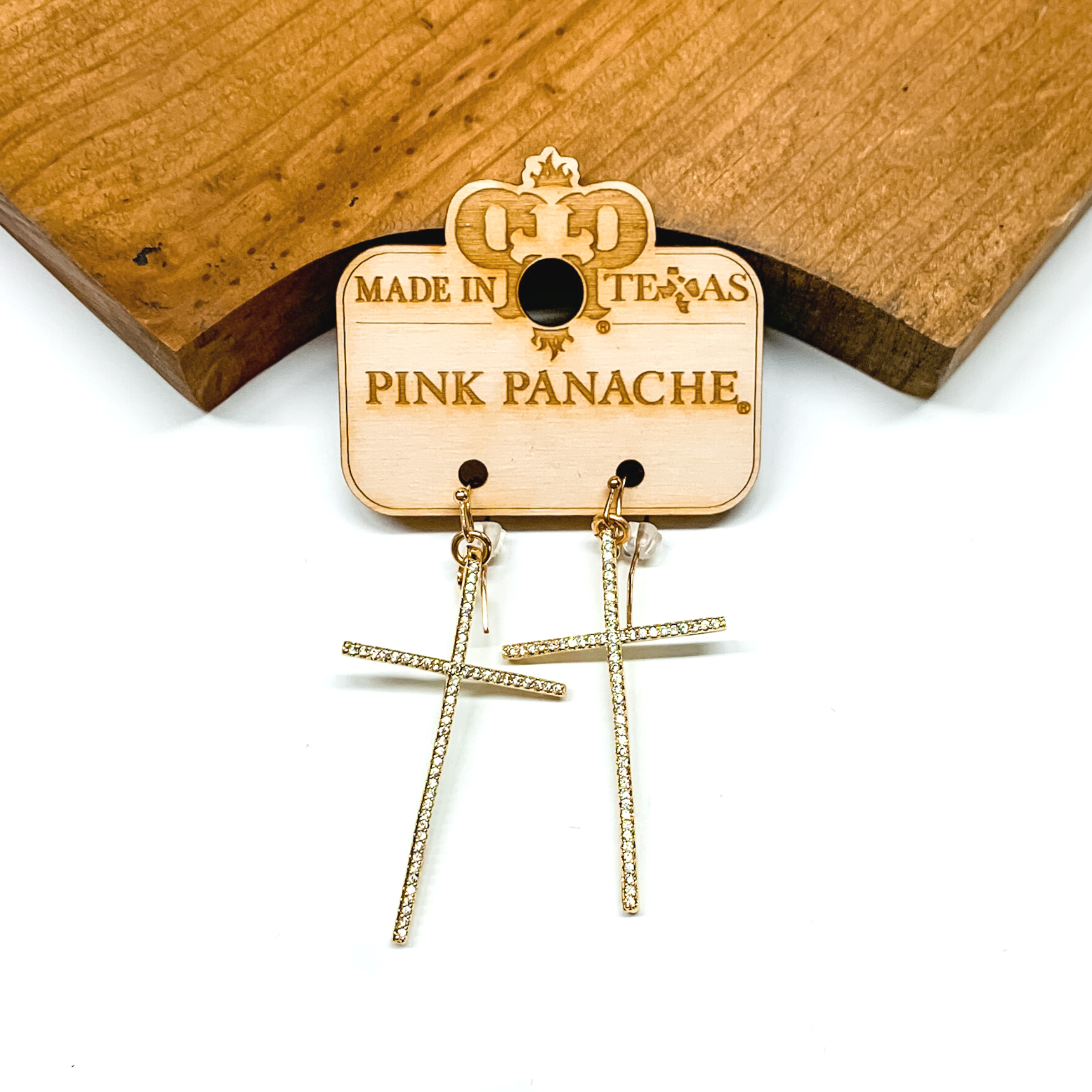 Gold cross earrings with an inlay of clear crystals. These earrings are pictured in front of a brown block on a white background. 