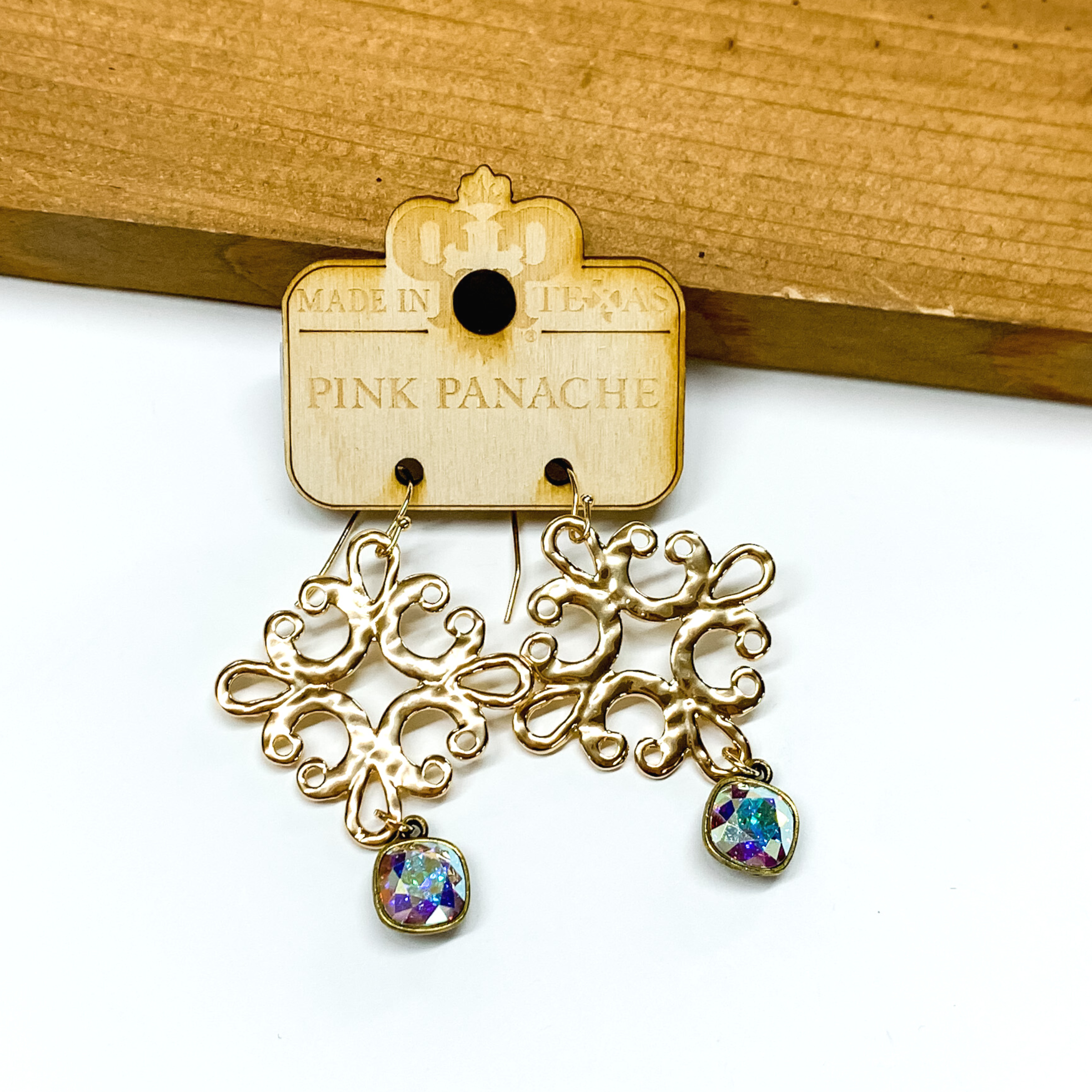 Hanging gold quatrefoil earrings with a hanging ab cushion crystal. these earrings are pictured in front of a brown block on a white background. 