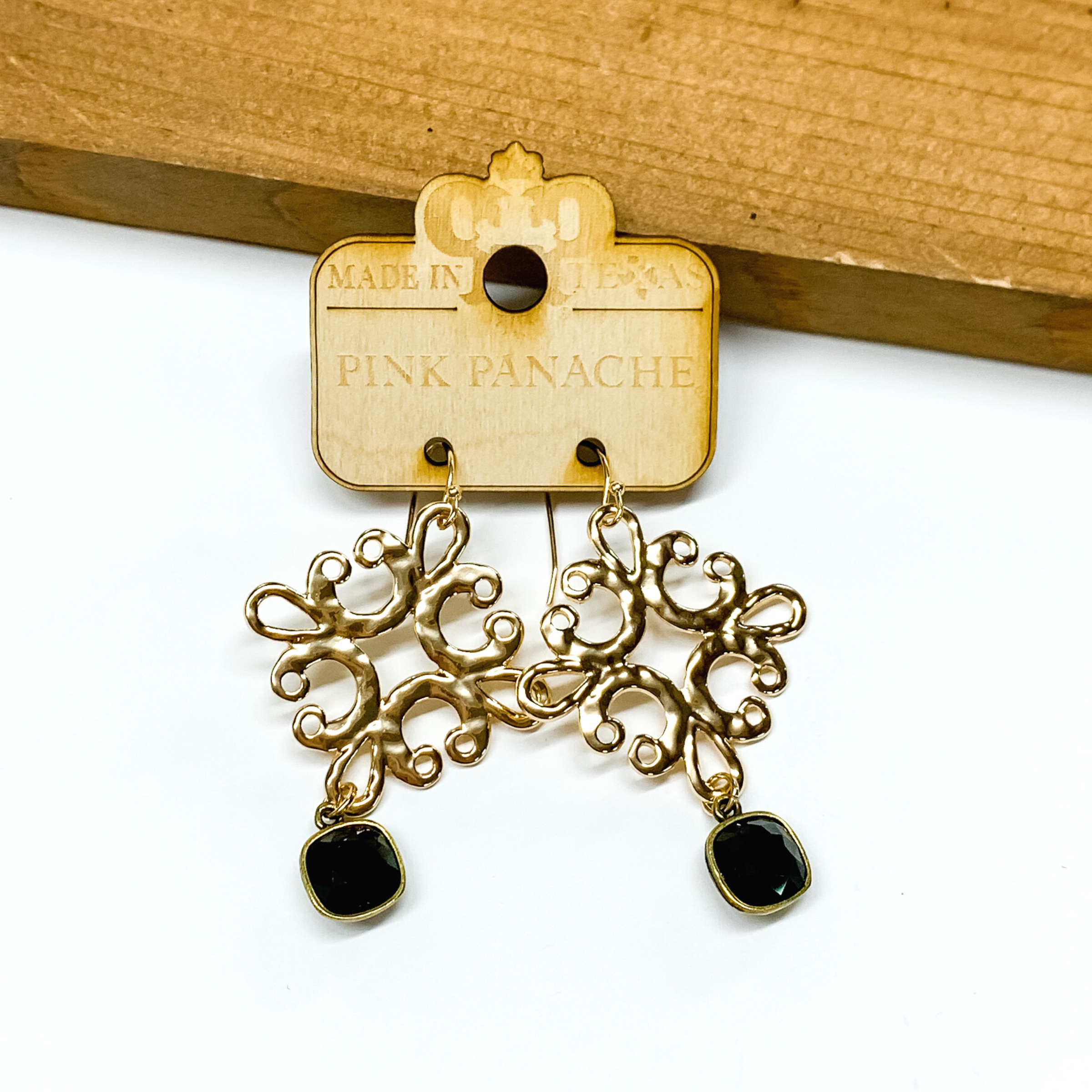 Hanging gold quatrefoil earrings with a hanging black cushion cut crystal. these earrings are pictured in front of a brown block on a white background. 