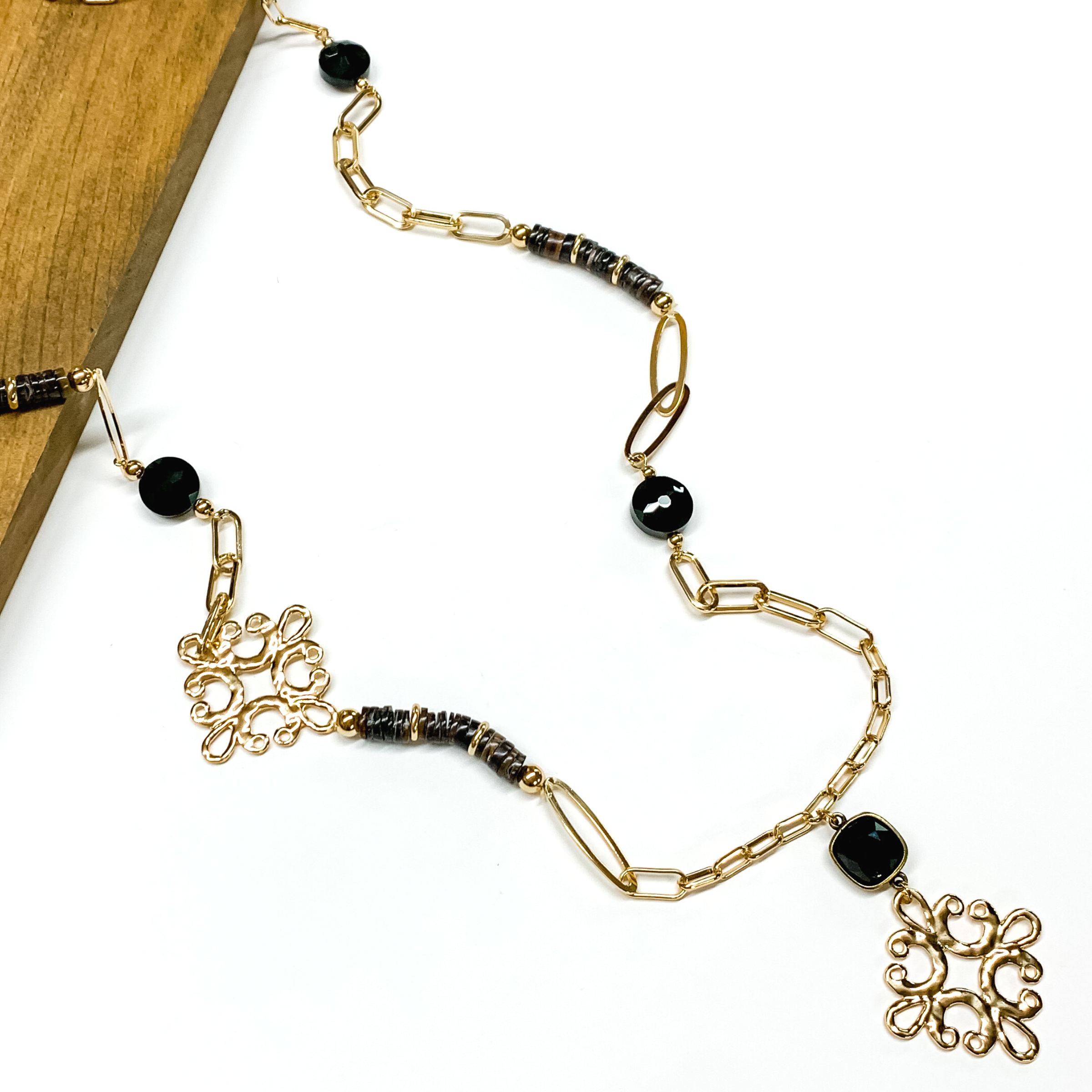 Gold paperclip chain and black beaded necklace. This necklace also includes a black crystal and gold quatrefoil drop. This necklace is pictured partially laying on a brown block on a white background. 