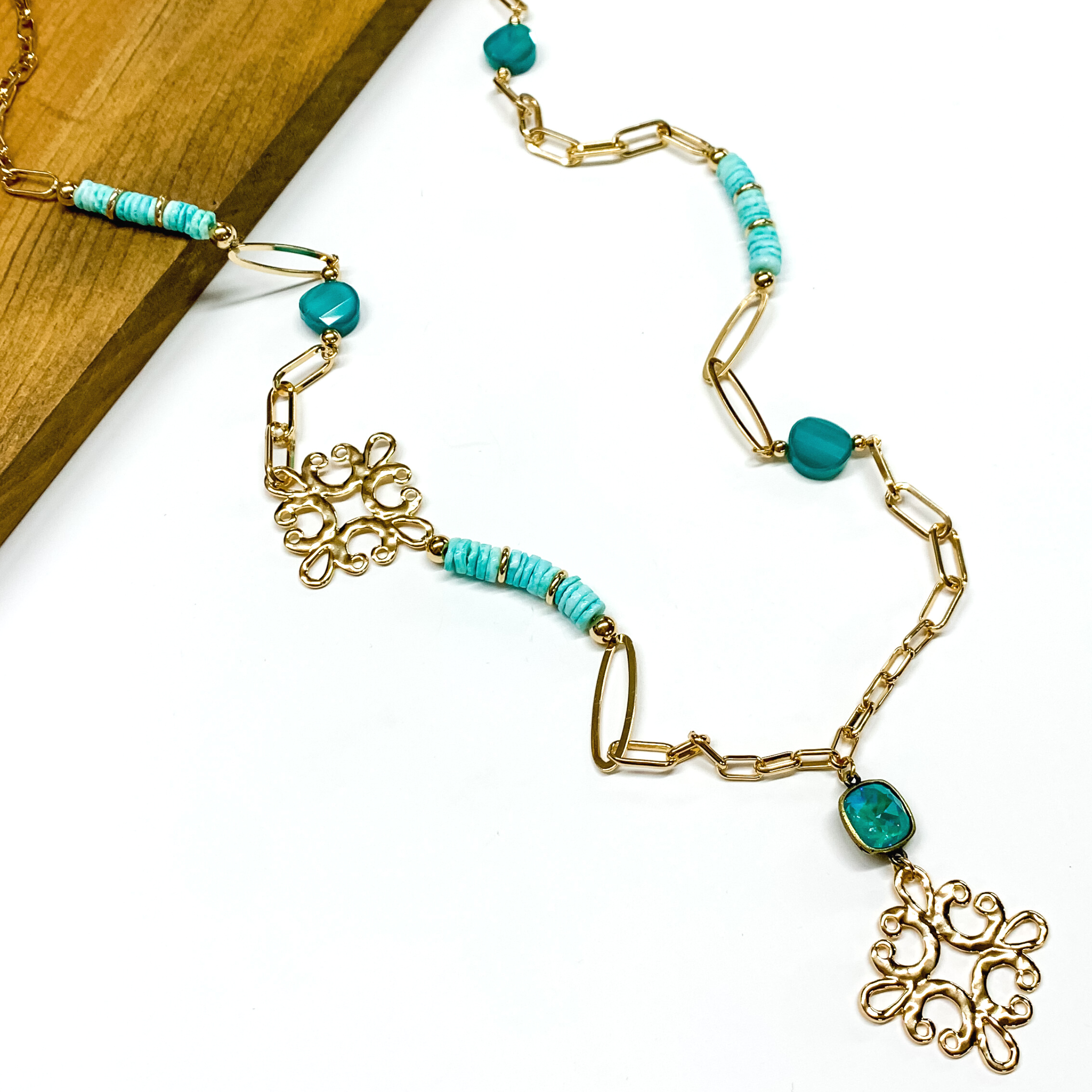 Gold paperclip chain and turqiose beaded necklace. This necklace also includes a turquoise crystal and gold quatrefoil drop. This necklace is pictured partially laying on a brown block on a white background. 