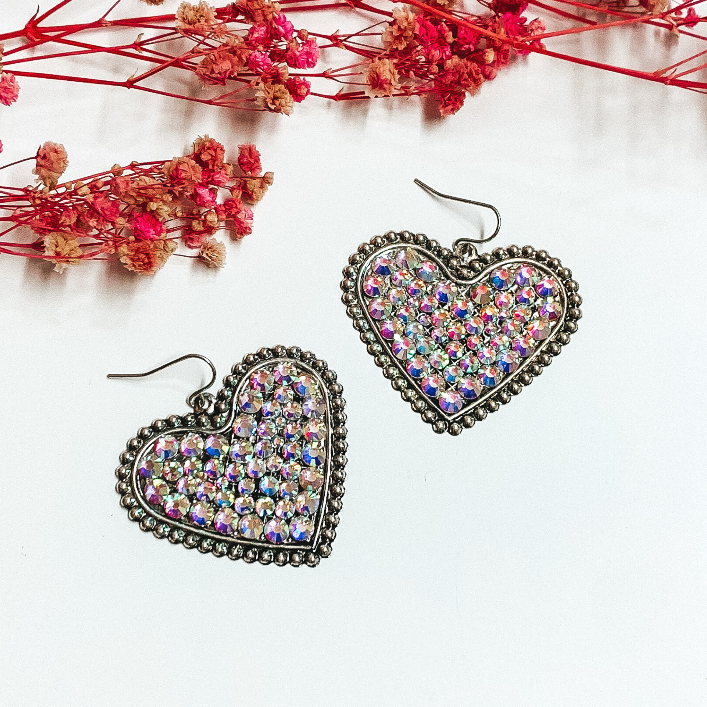 Silver heart dangle earrings encrusted with ab crystals. These earrings are pictured on a white background with small pink flowers at the top of the picture. 