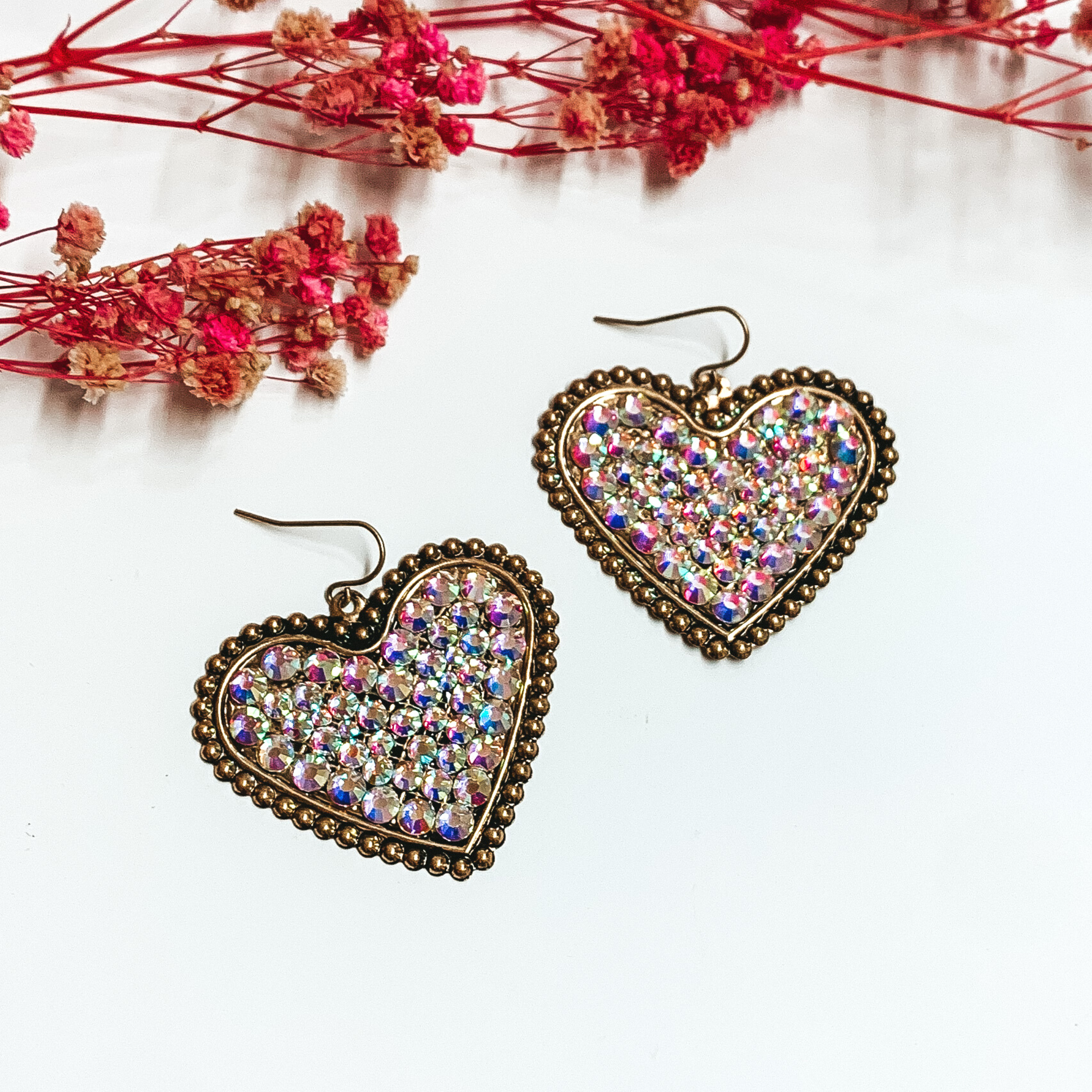 Bronze heart dangle earrings encrusted with ab crystals. These earrings are pictured on a white background with small pink flowers at the top of the picture. 
