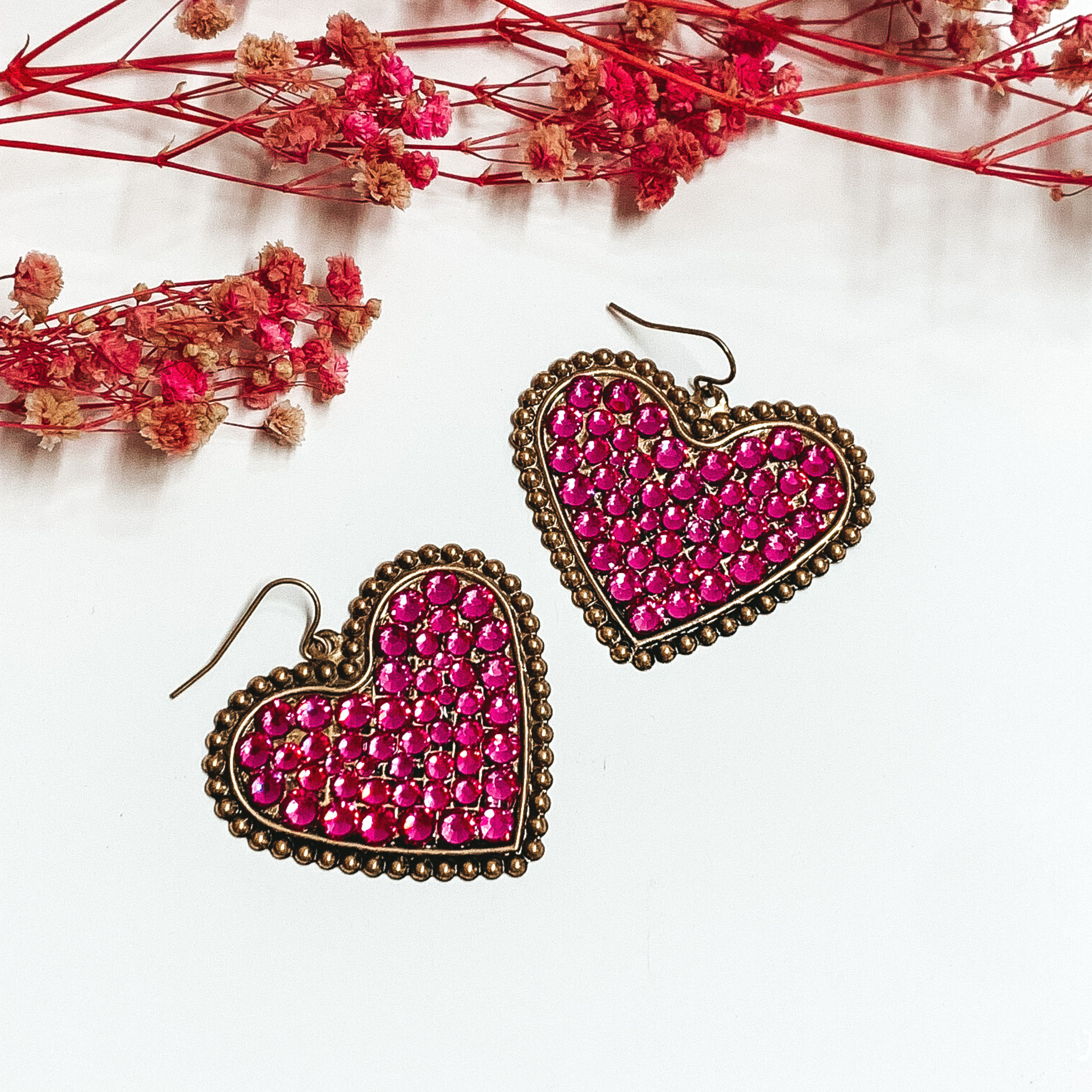 Bronze heart dangle earrings encrusted with fuchsia crystals. These earrings are pictured on a white background with small pink flowers at the top of the picture. 