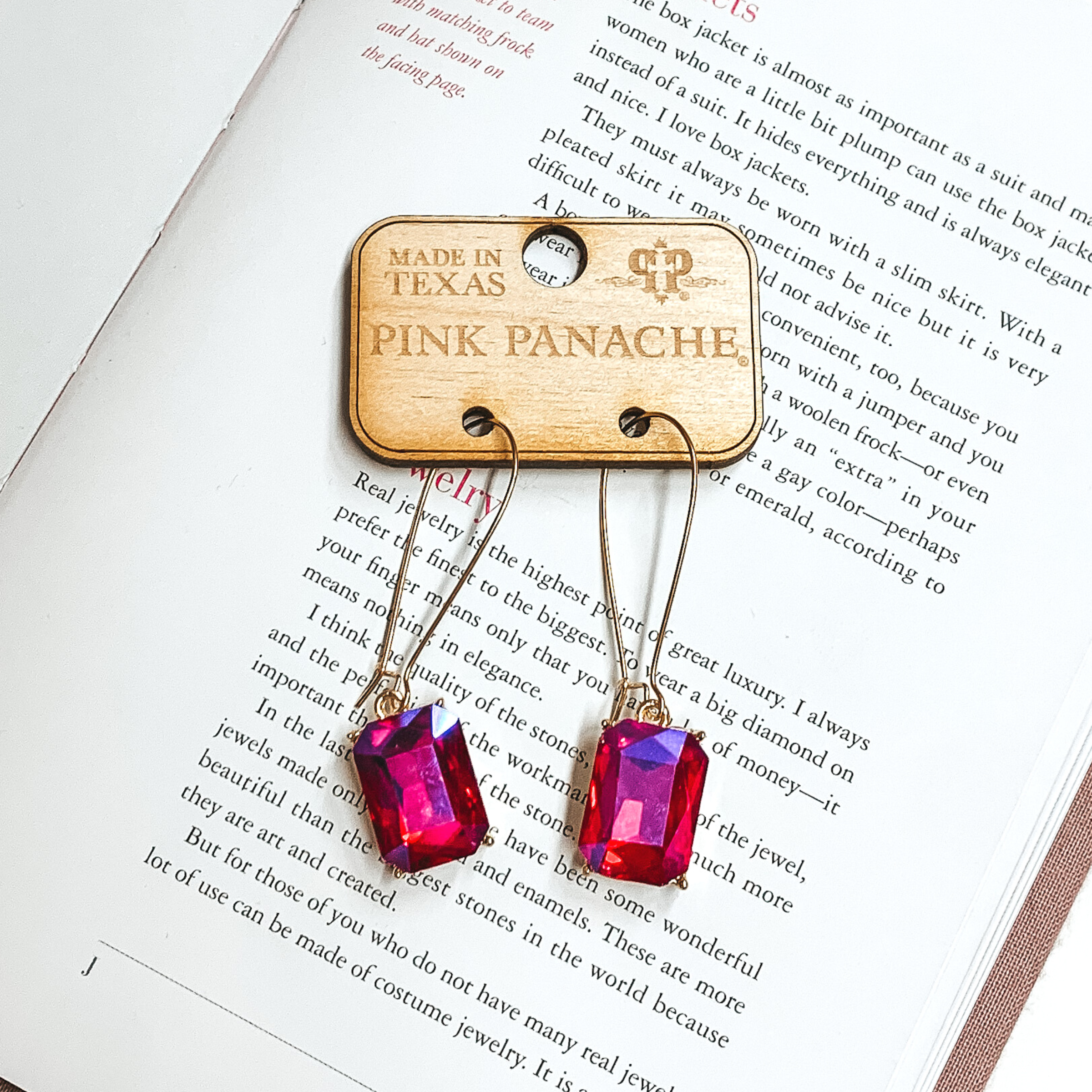Thin, gold wire earrings with a rectangle, crystal drop. This crystal is a red ab color. This pair of earrings is pictured on an open book. 