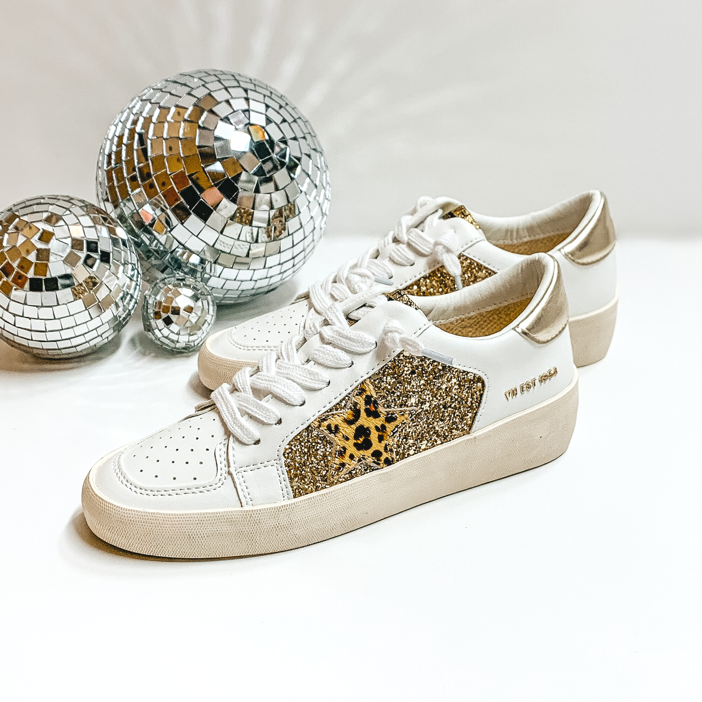White tennis shoes with a patch of white gold glitter and a leopard print star emblem. These shoes also have white laces and a gold part on the heel. These shoes are pictured on a white background with disco balls on the left hand side.
