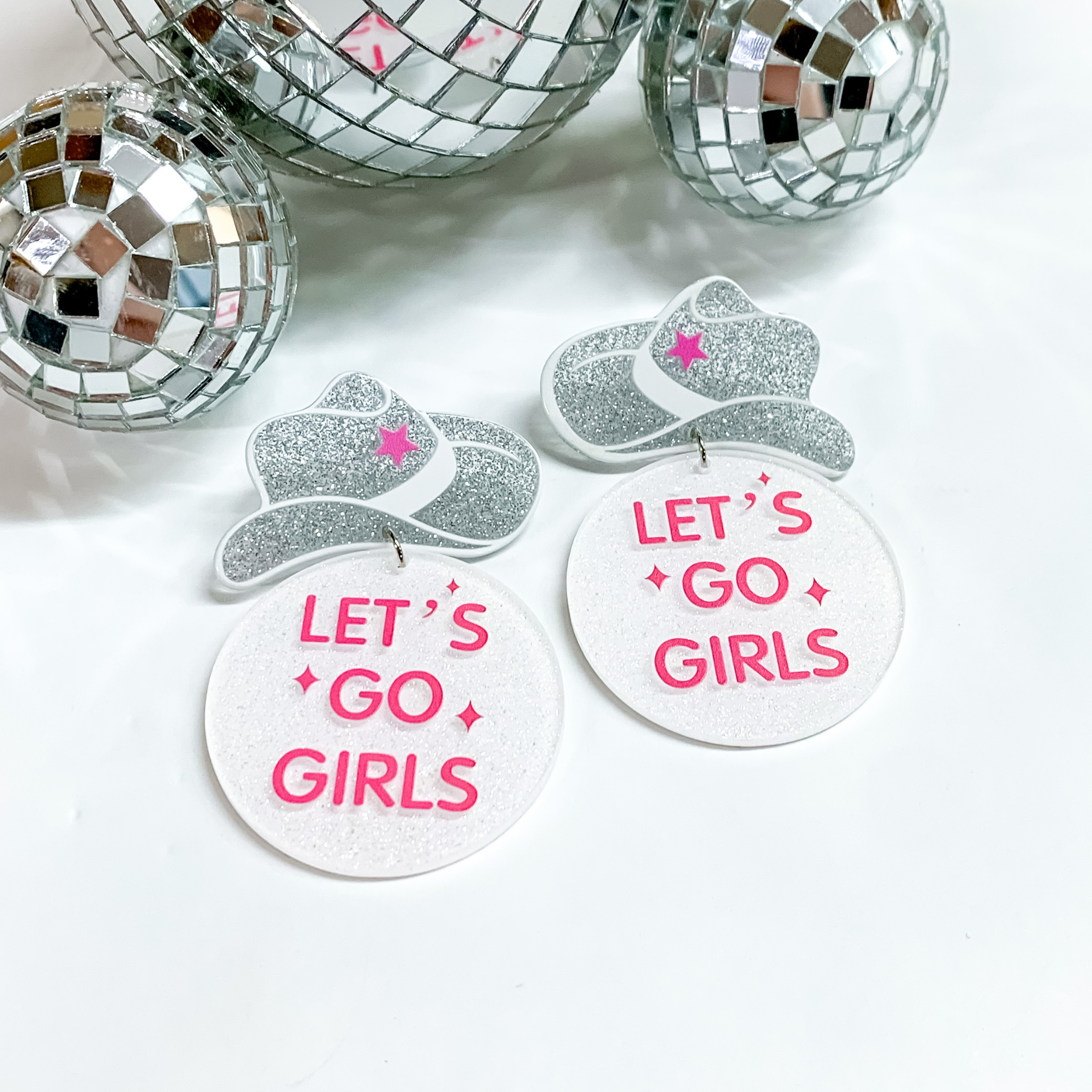 Silver glitter cowboy hat earrings with a white glitter circle drop. The saying "LET'S GO GIRLS" is printed in pink on the circle dorp. These earring are pictured on a white background with disco balls at the top of the picture. 