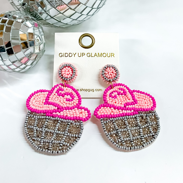 Beaded cowgirl hat and disco ball drop earrings. The hat is a pink color and the disco ball is silver. These earring are pictured on a white background with disco balls at the top of the picture. 