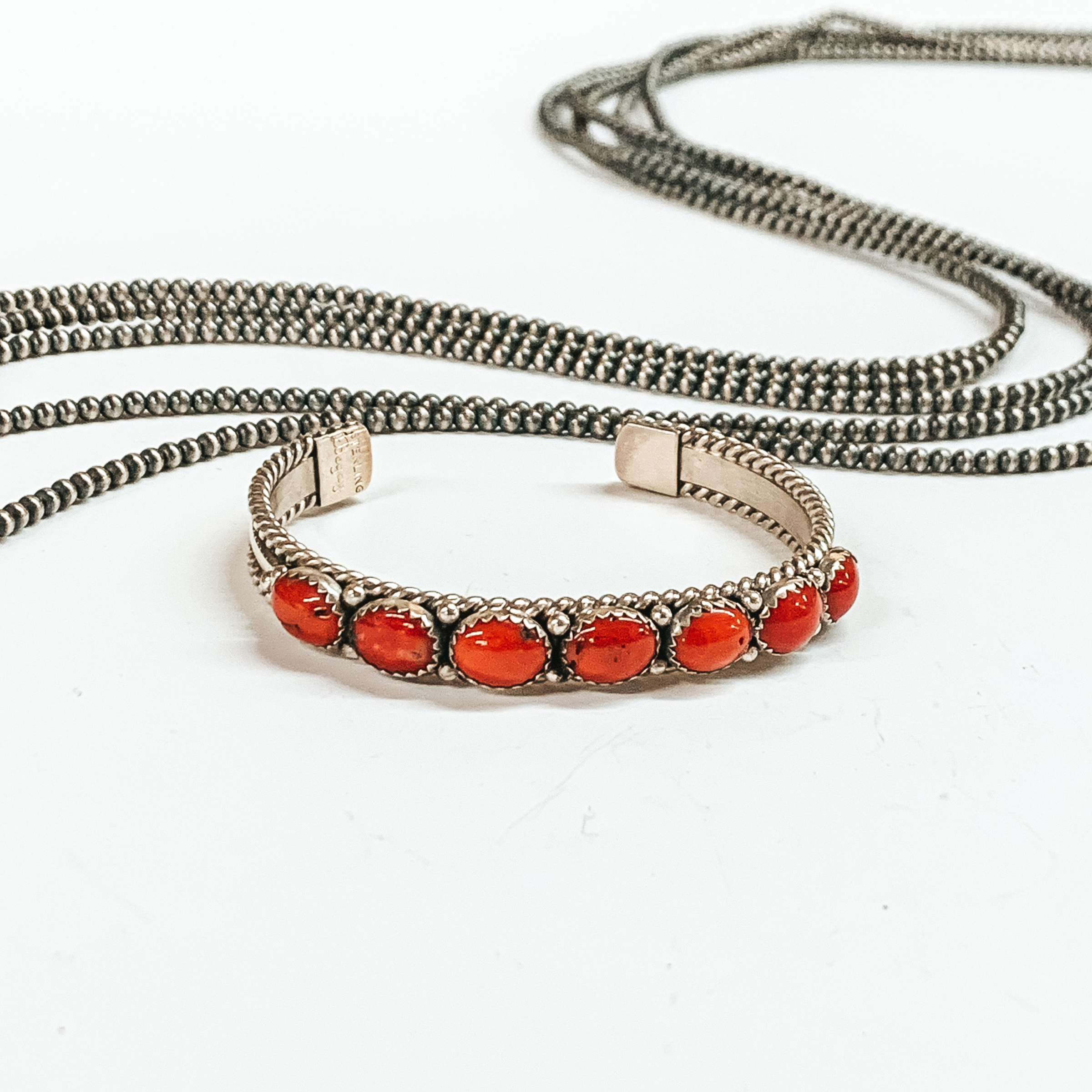 Silver cuff bracelet with a rope detailed outline. This bracelet also includes seven red coral, oval stones. This bracelet is pictured on a white background with silver beads behind it. 