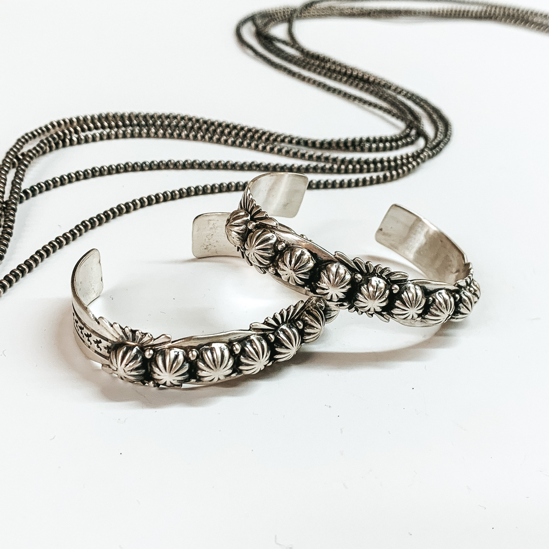 Two silver cuff bracelet with engraving details. This bracelet is pictured on a white background with silver beads behind it. 
