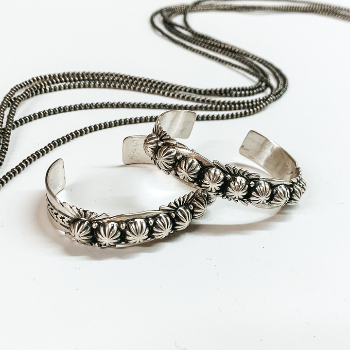 Two silver cuff bracelet with engraving details. This bracelet is pictured on a white background with silver beads behind it. 