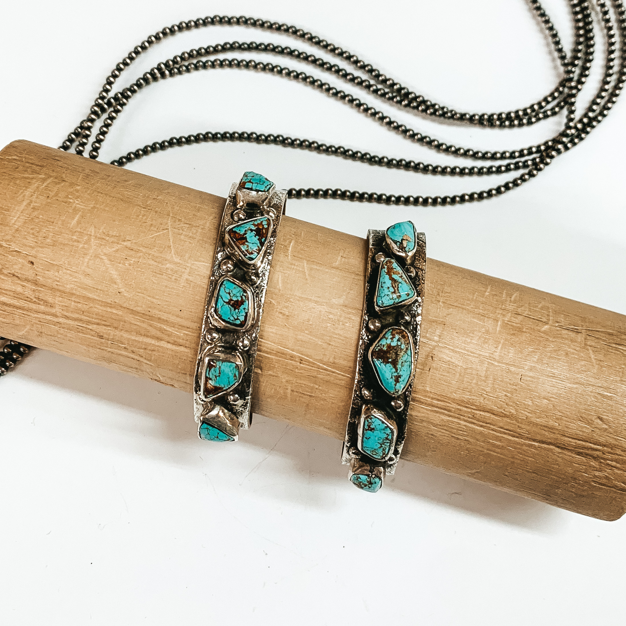 Two silver bracelets with engraving details and irregular shaped turquoise stones. This bracelet is pictured on a on a wooden bracelet holder on a white background with silver beads behind it. 