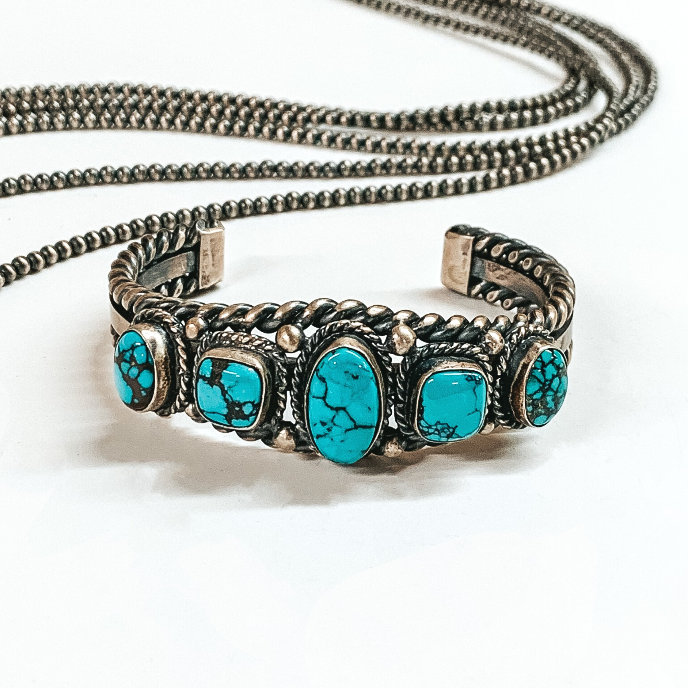 Silver cuff bracelet with a twisted rope outline and five turquoise stones. These bracelets are pictured on a white background with silver beads behind it. 