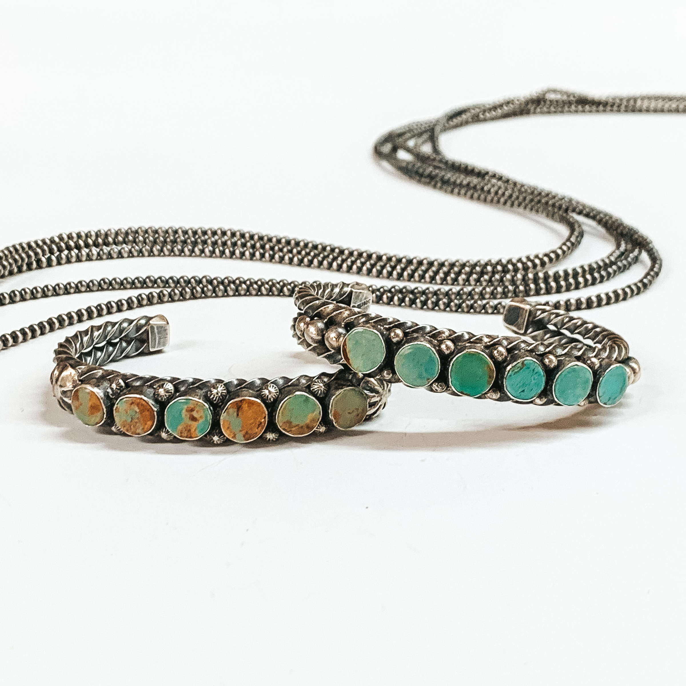 Two silver twisted bracelets with six turquoise stones. These bracelets are pictured on a white background with silver beads behind it. 