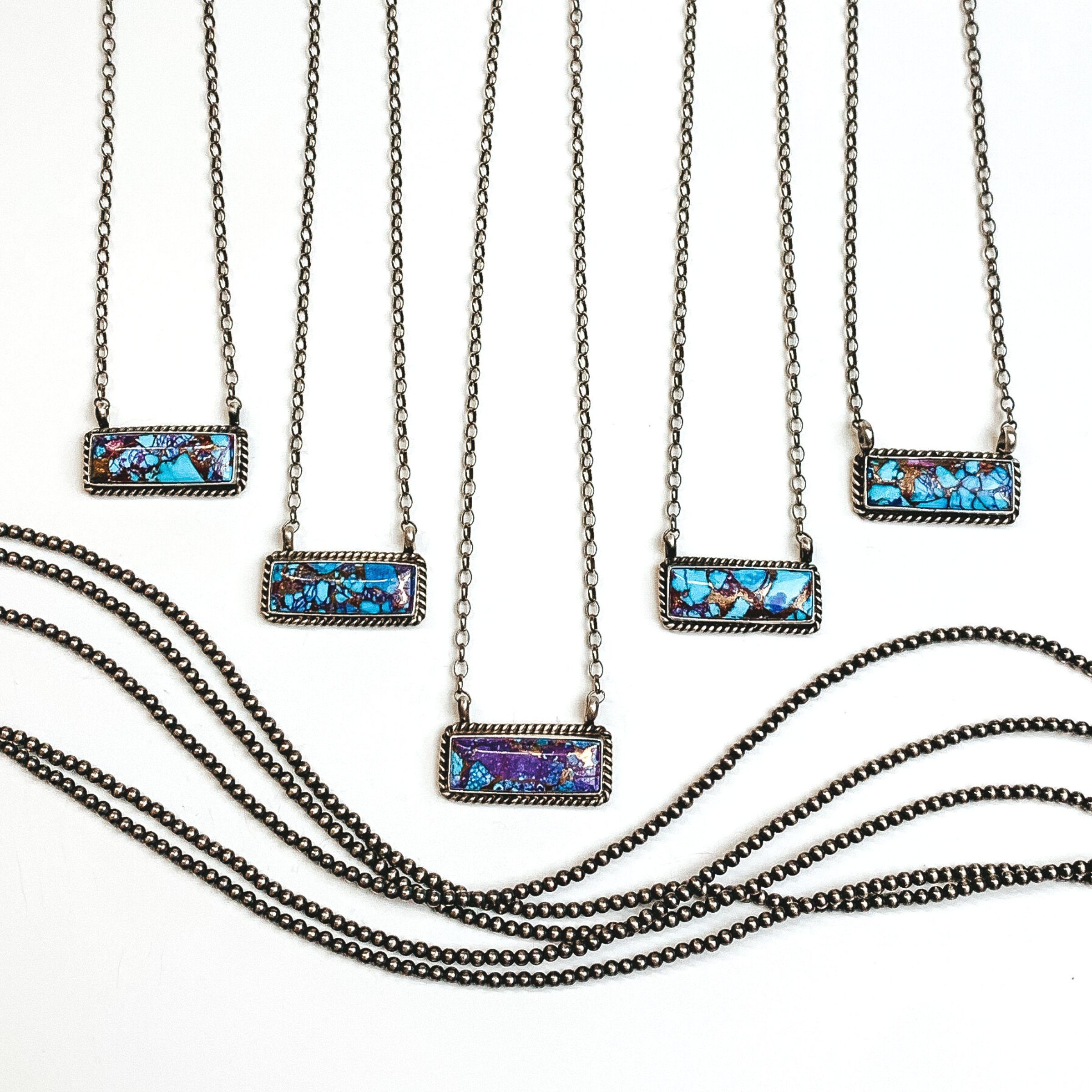 Four silver chain necklaces with rectangle bars that have a purple mojave and turquoise remix stone. These necklaces are pictured on a white background with silver beads under the necklaces. 