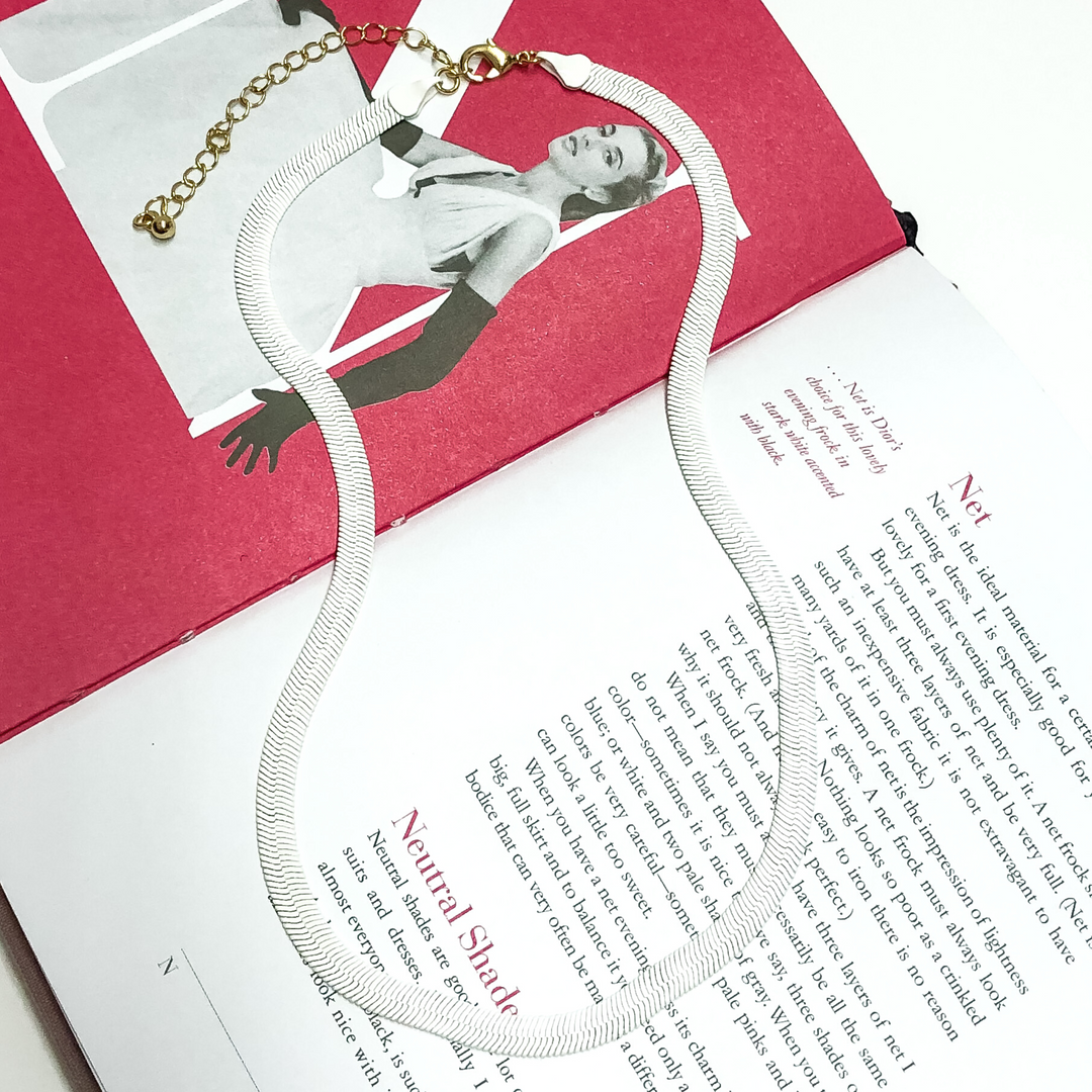 This is a white herringbone chain necklace, with a gold extender. It is on a open book, with a white background.